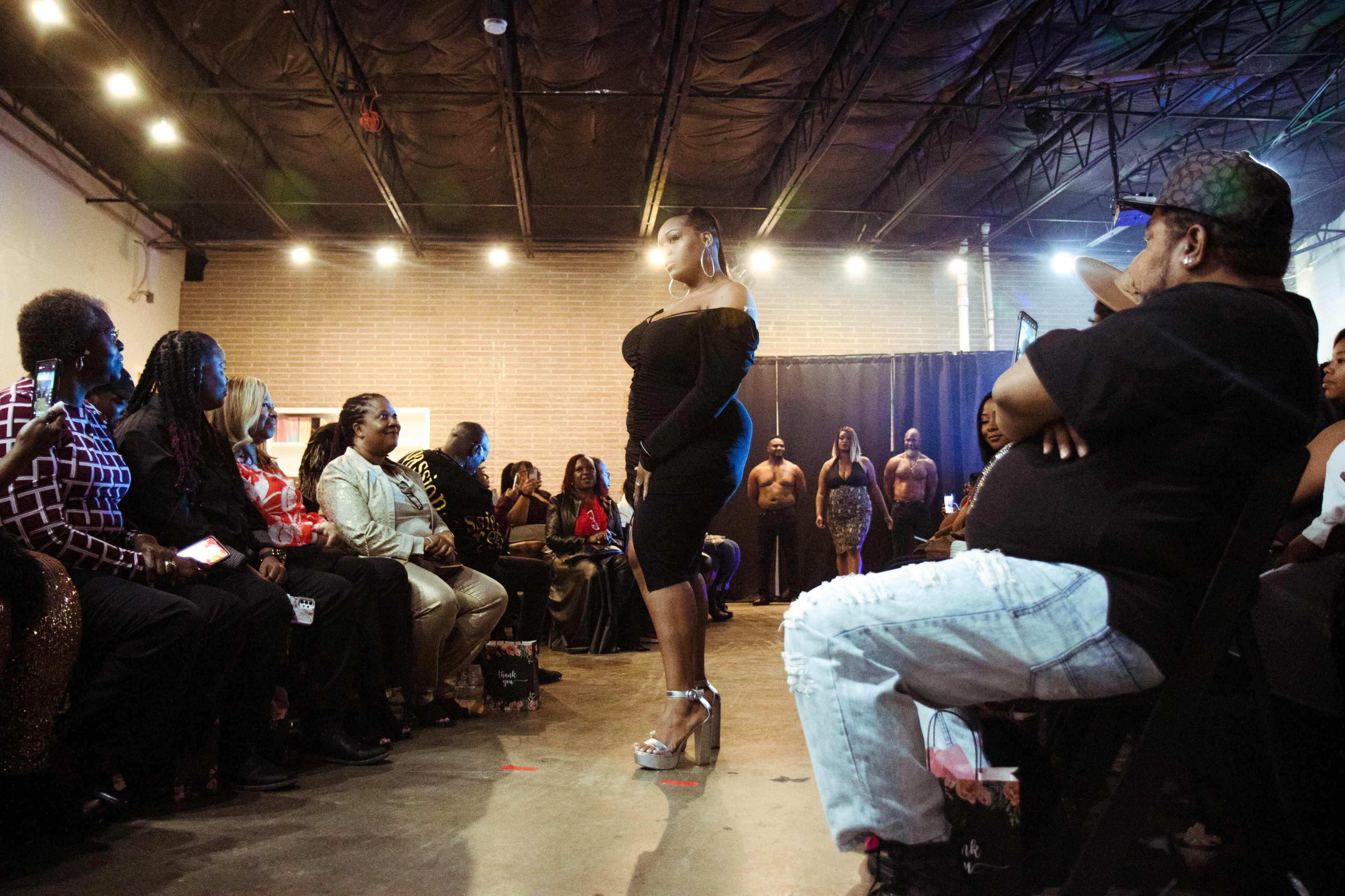 A model walks down a runway in front of an audience seated on both sides, with several people in formal attire watching attentively.