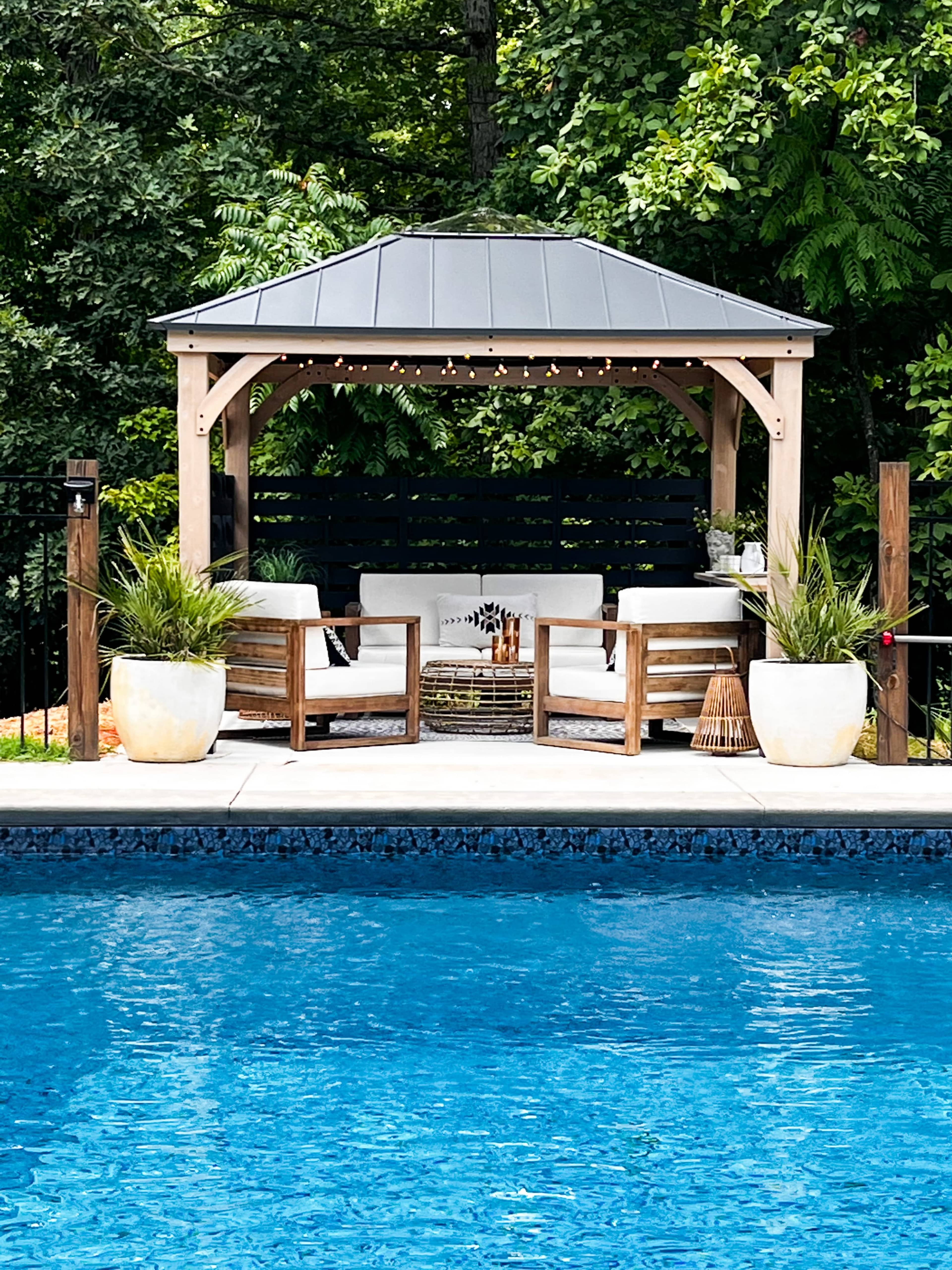 A wooden gazebo with seating is positioned beside a swimming pool, surrounded by lush greenery.