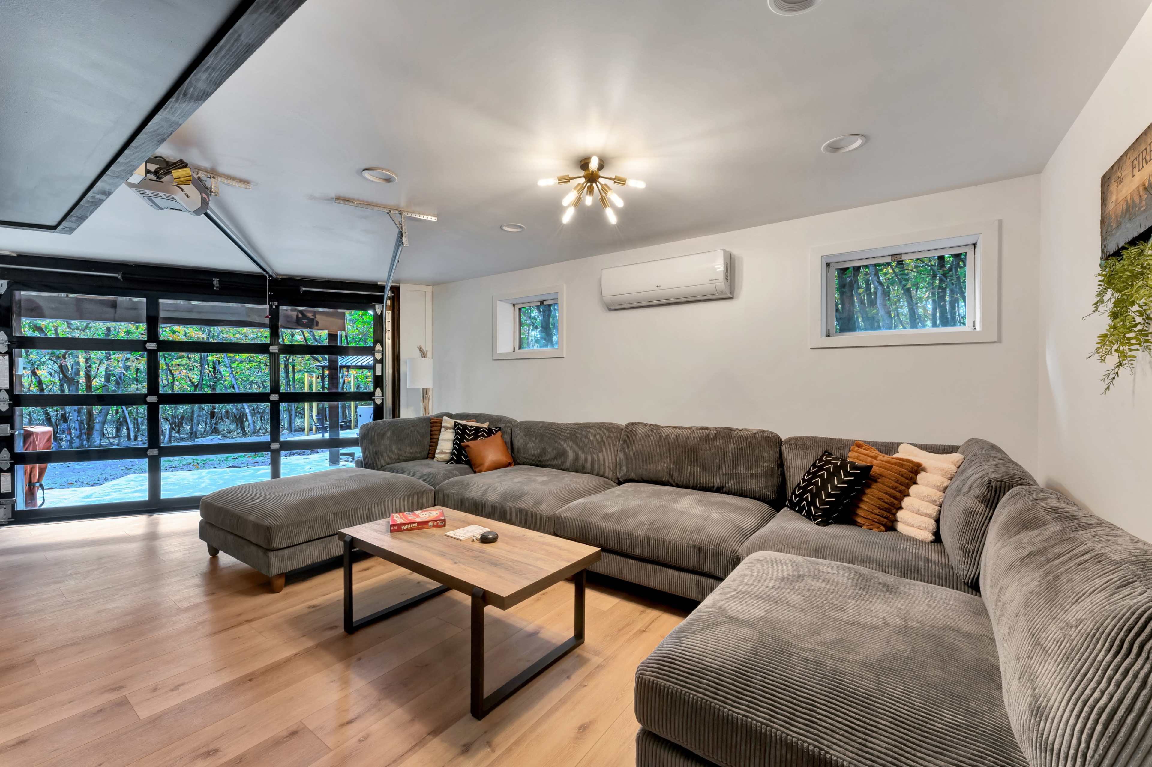 The image shows a modern living room with a large gray sectional sofa, a wooden coffee table, and a glass garage door that opens to a pool area.