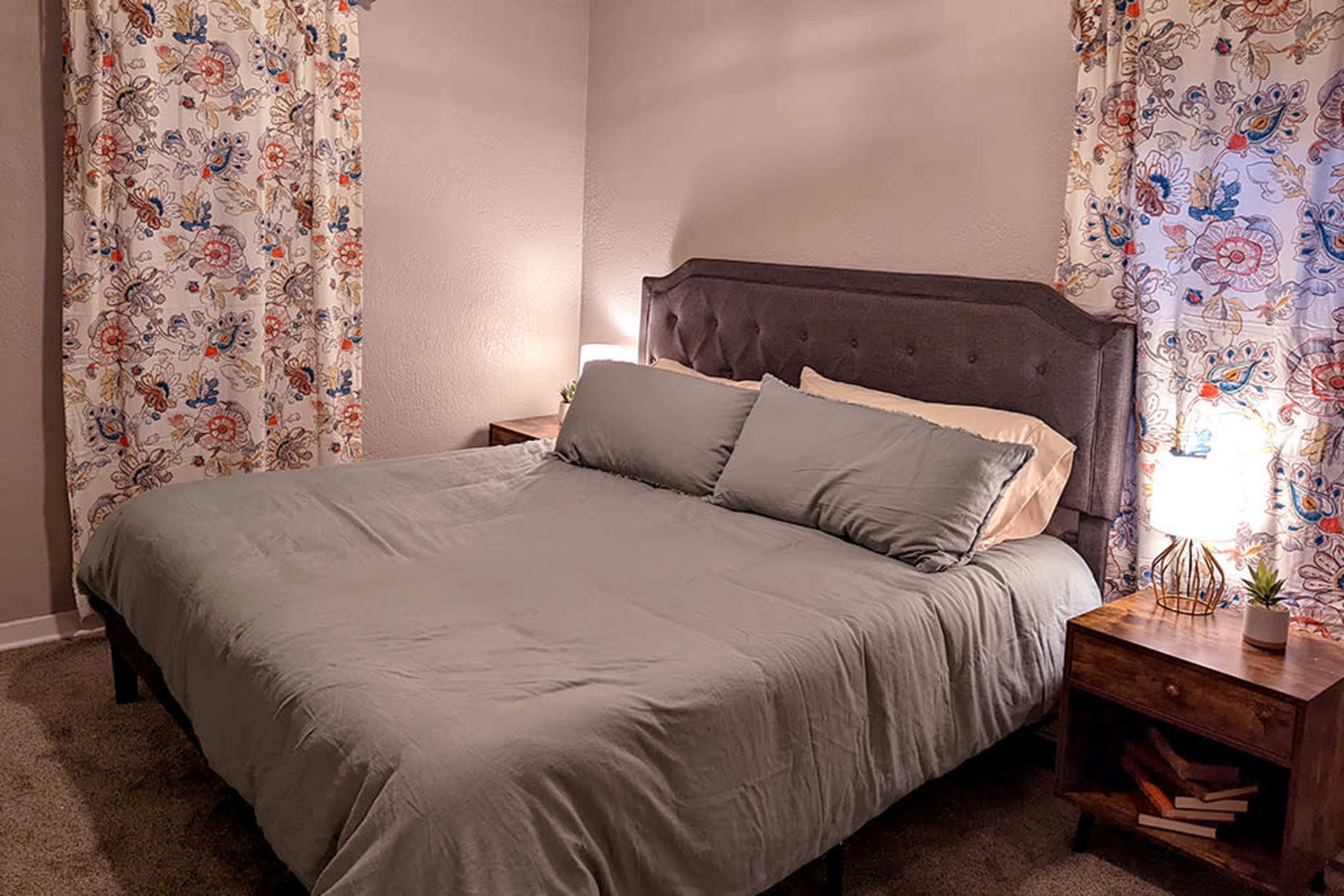 A neatly arranged bedroom with a gray bedspread, two pillows, and decorative curtains with floral patterns, beside wooden nightstands and lamps.