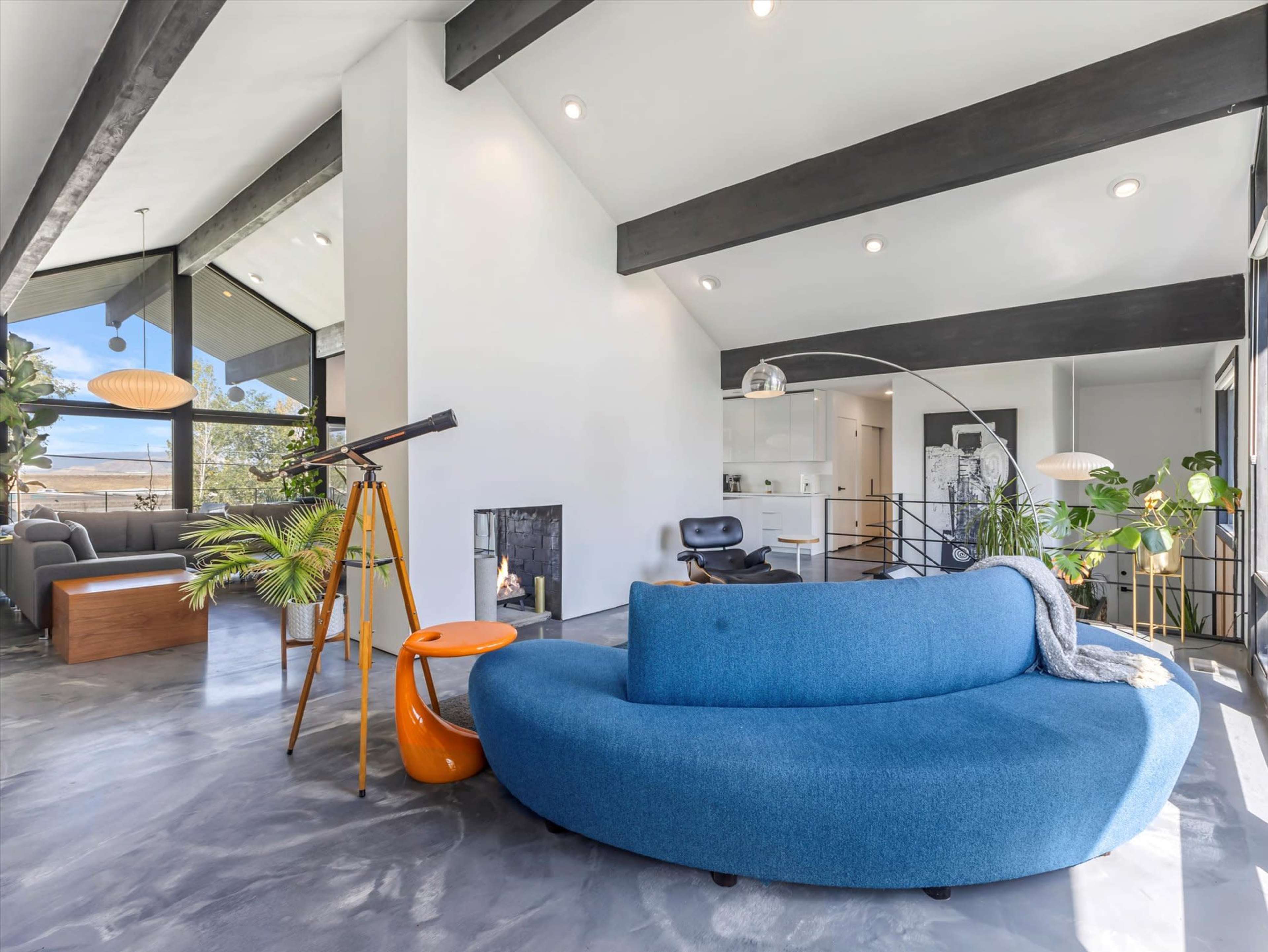 The image shows a modern living space with a blue circular sofa, a telescope, and large windows that allow natural light to enter.