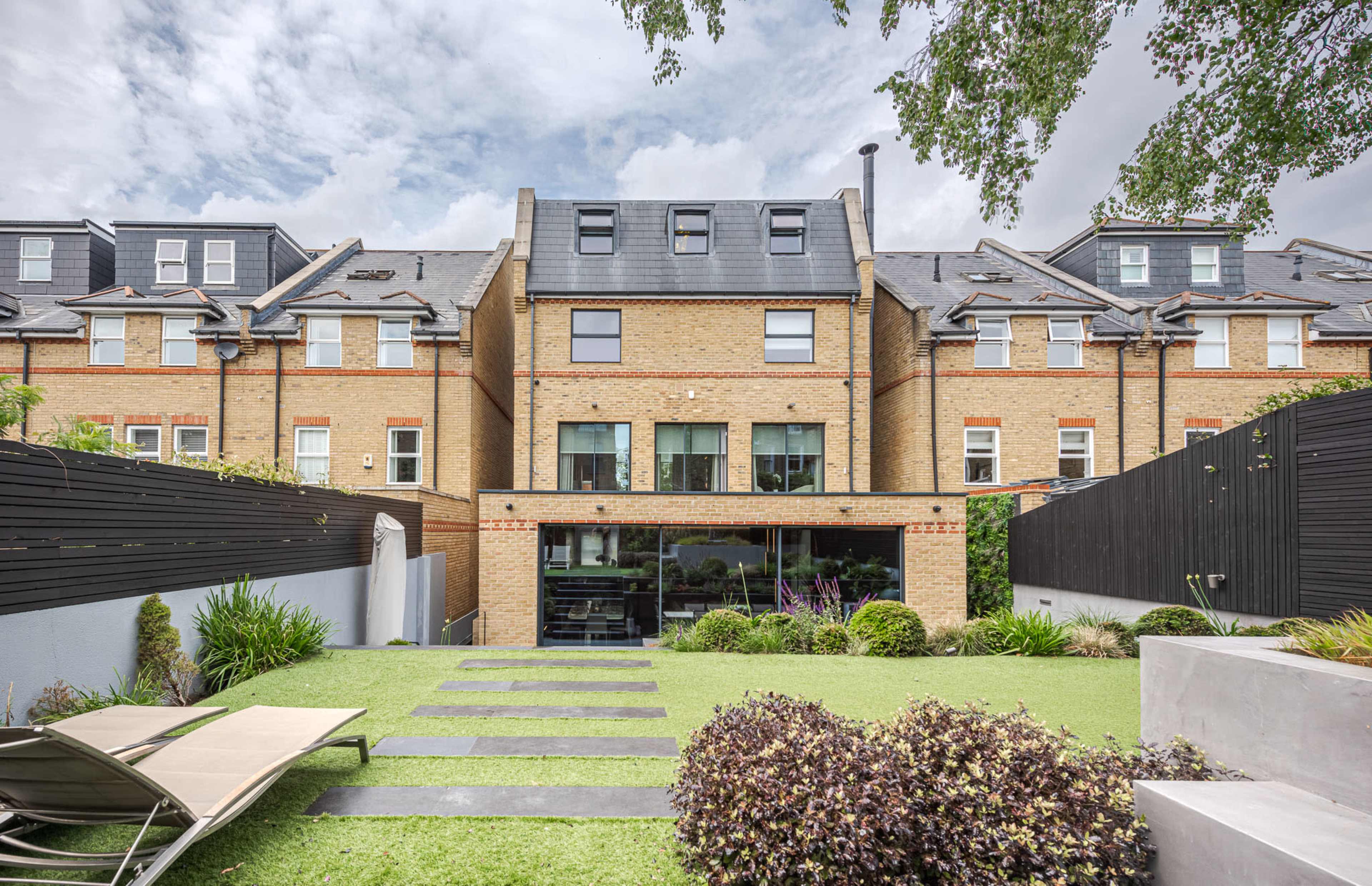 The image shows a modern brick townhouse with large windows, surrounded by a neatly landscaped garden featuring a grassy area and stone paths.