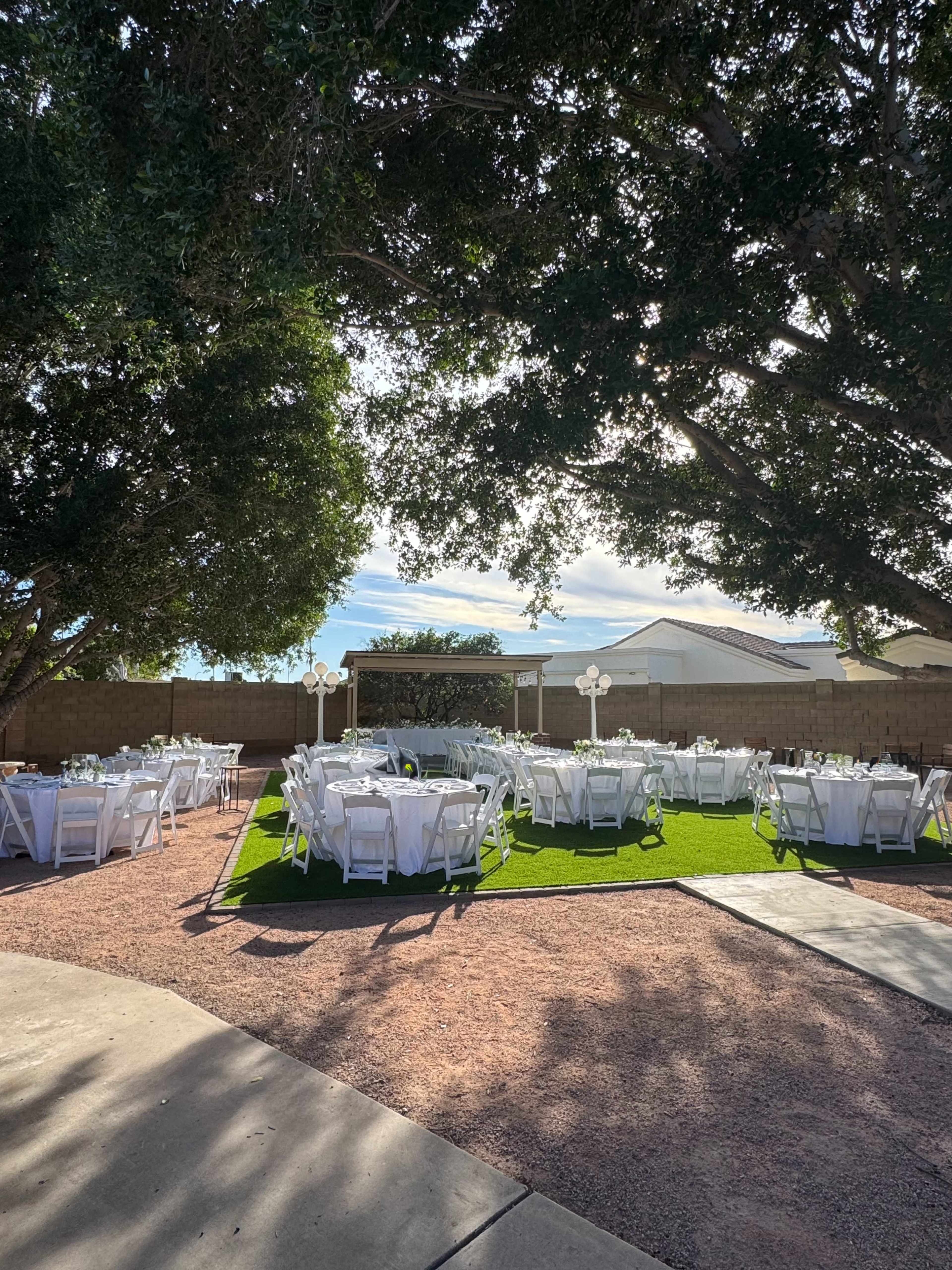 A landscaped outdoor event space features several round tables with white tablecloths arranged on green grass beneath large trees.