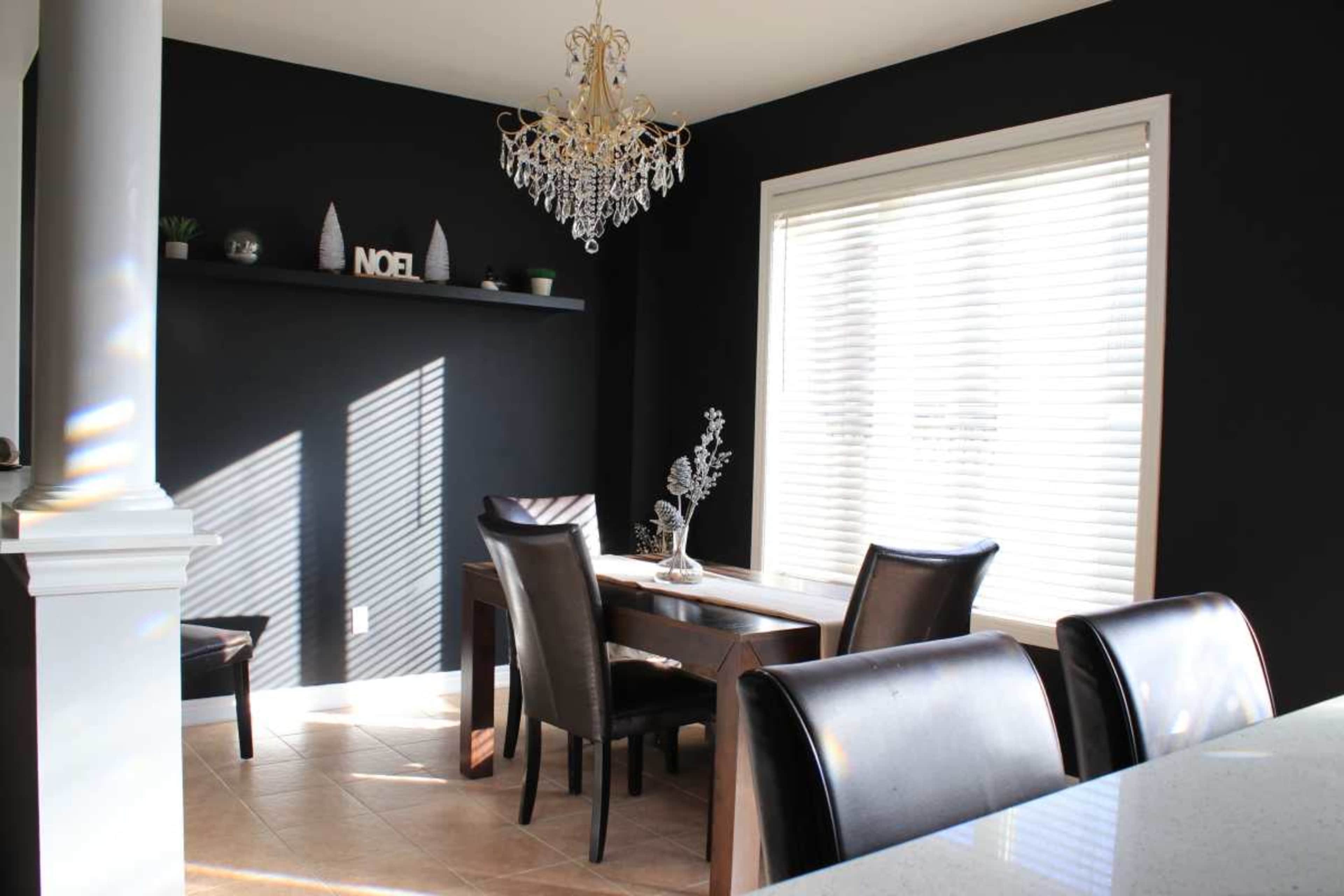 A dining area features a wooden table with chairs beneath a chandelier and a shelf decorated with small items against a dark wall.