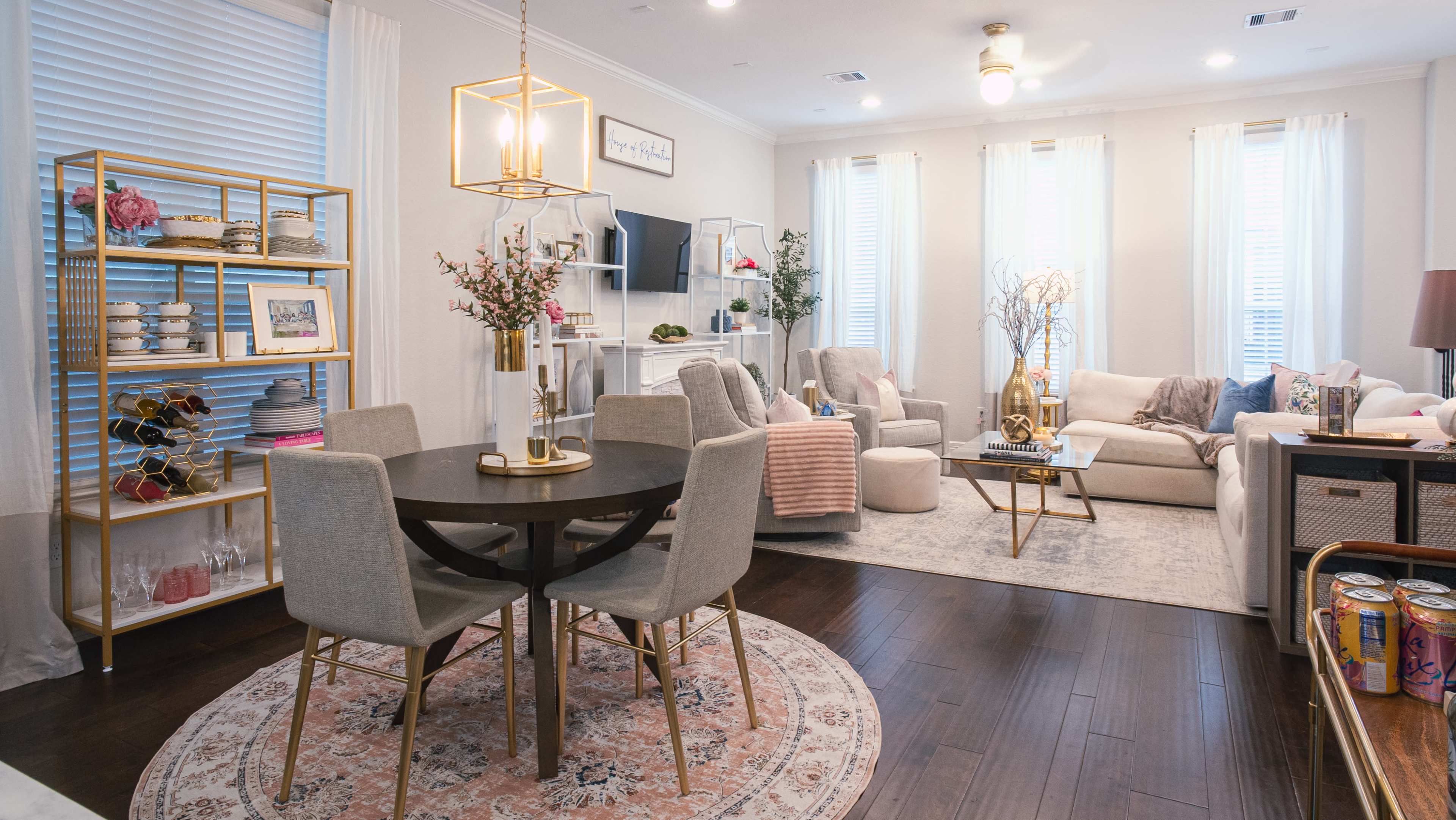 A well-furnished living room and dining area featuring a round table with gray chairs, a light-colored sofa, decorative shelves, and large windows with sheer curtains.