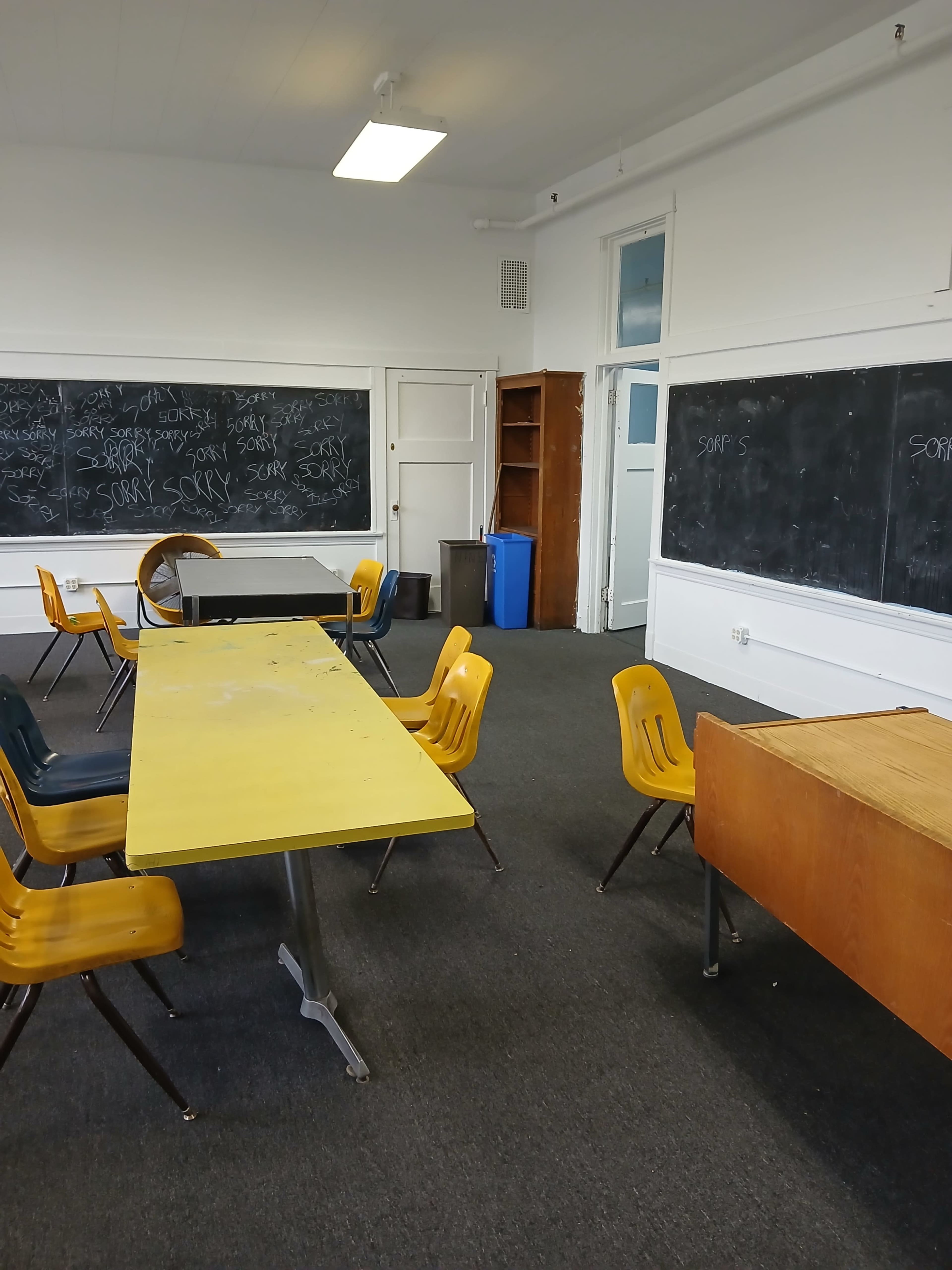 The image shows an empty classroom with yellow tables and chairs, black chalkboards covered in writing, and a storage cabinet in the corner.