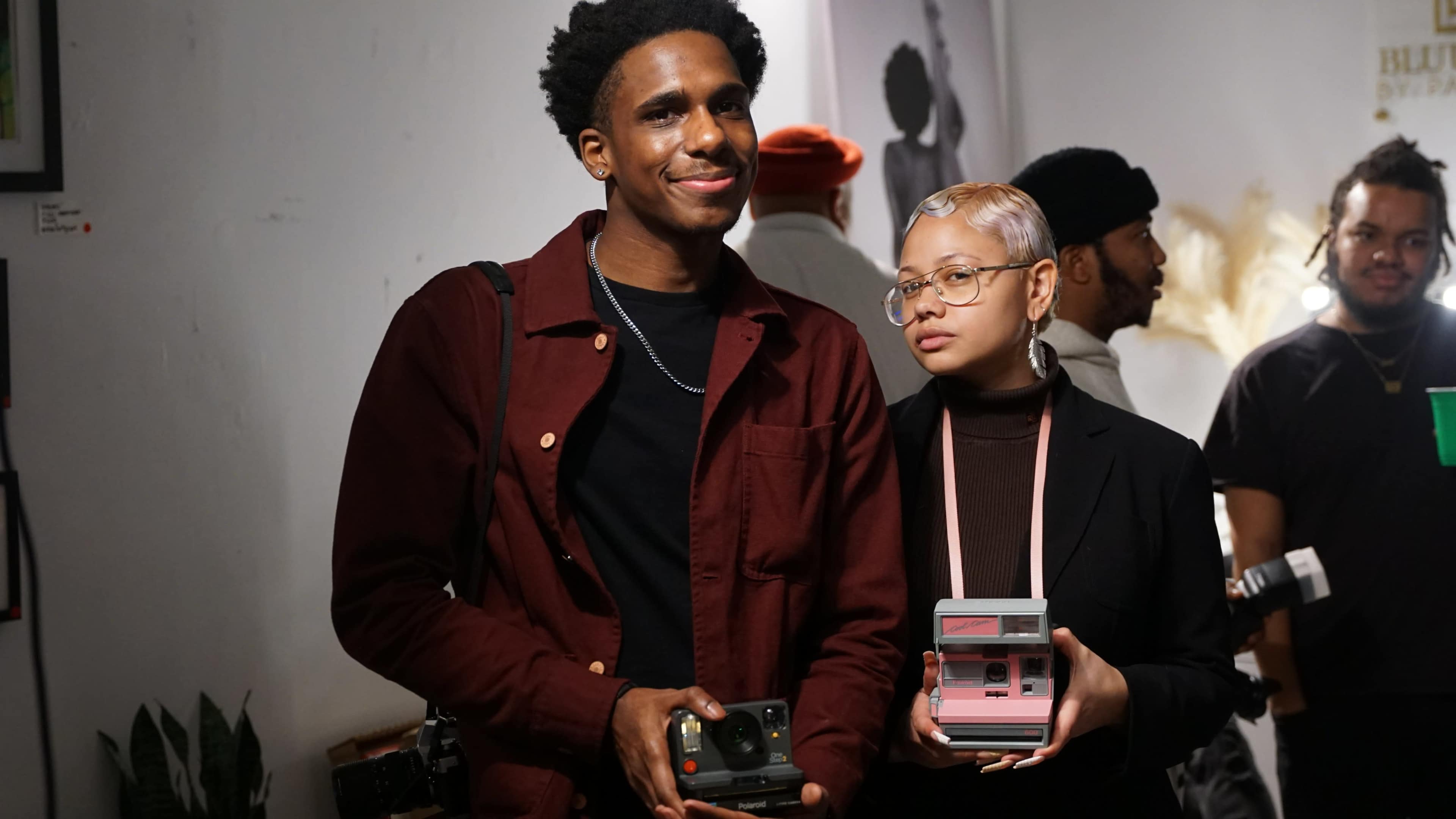 A man and a woman pose together, each holding a camera, in an art gallery setting with various artworks displayed in the background.