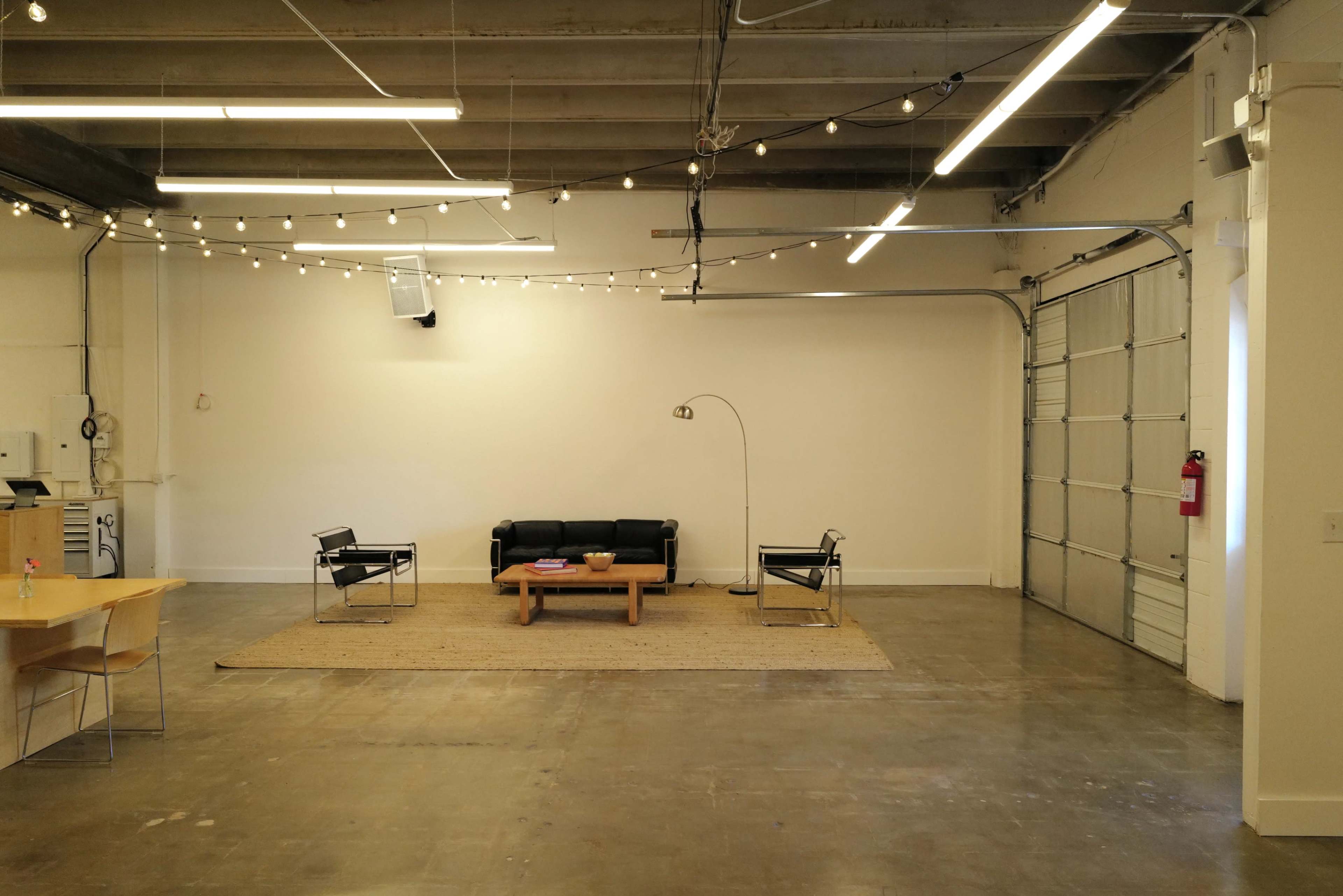 A spacious, minimalist room features a black sofa and two chairs arranged around a coffee table on a rug, with string lights overhead and a large garage door to one side.