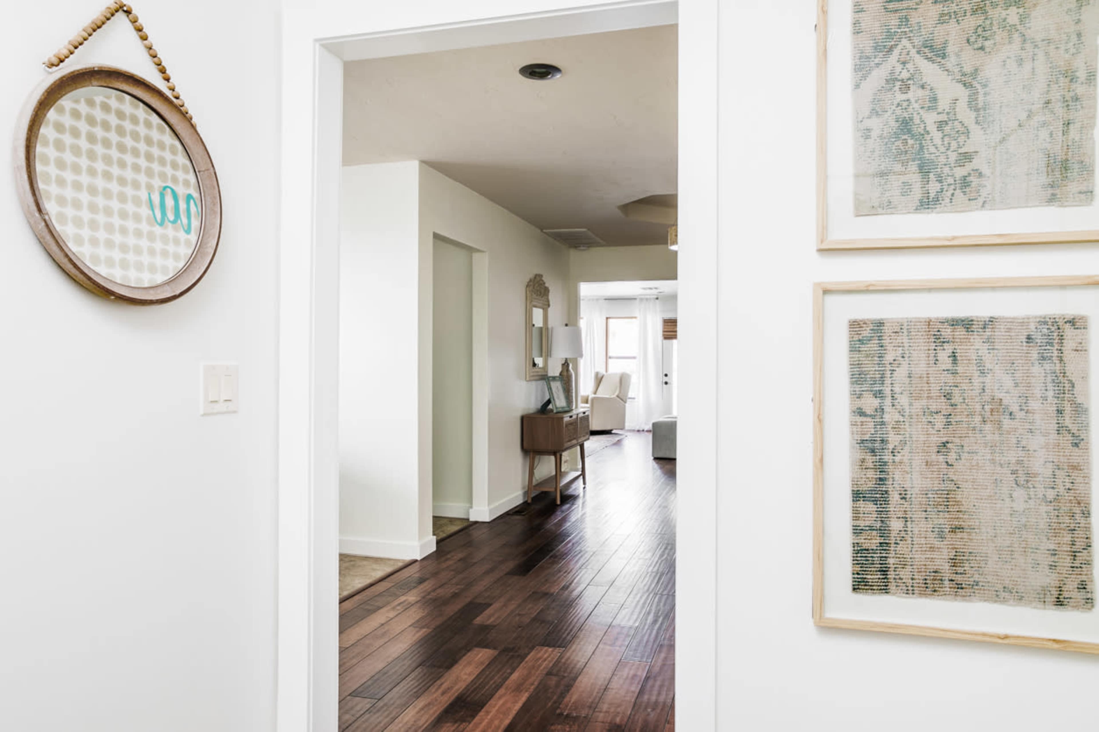 A narrow hallway leads to a room with light-colored furniture and wooden flooring, framed by two wall hangings and a round mirror.