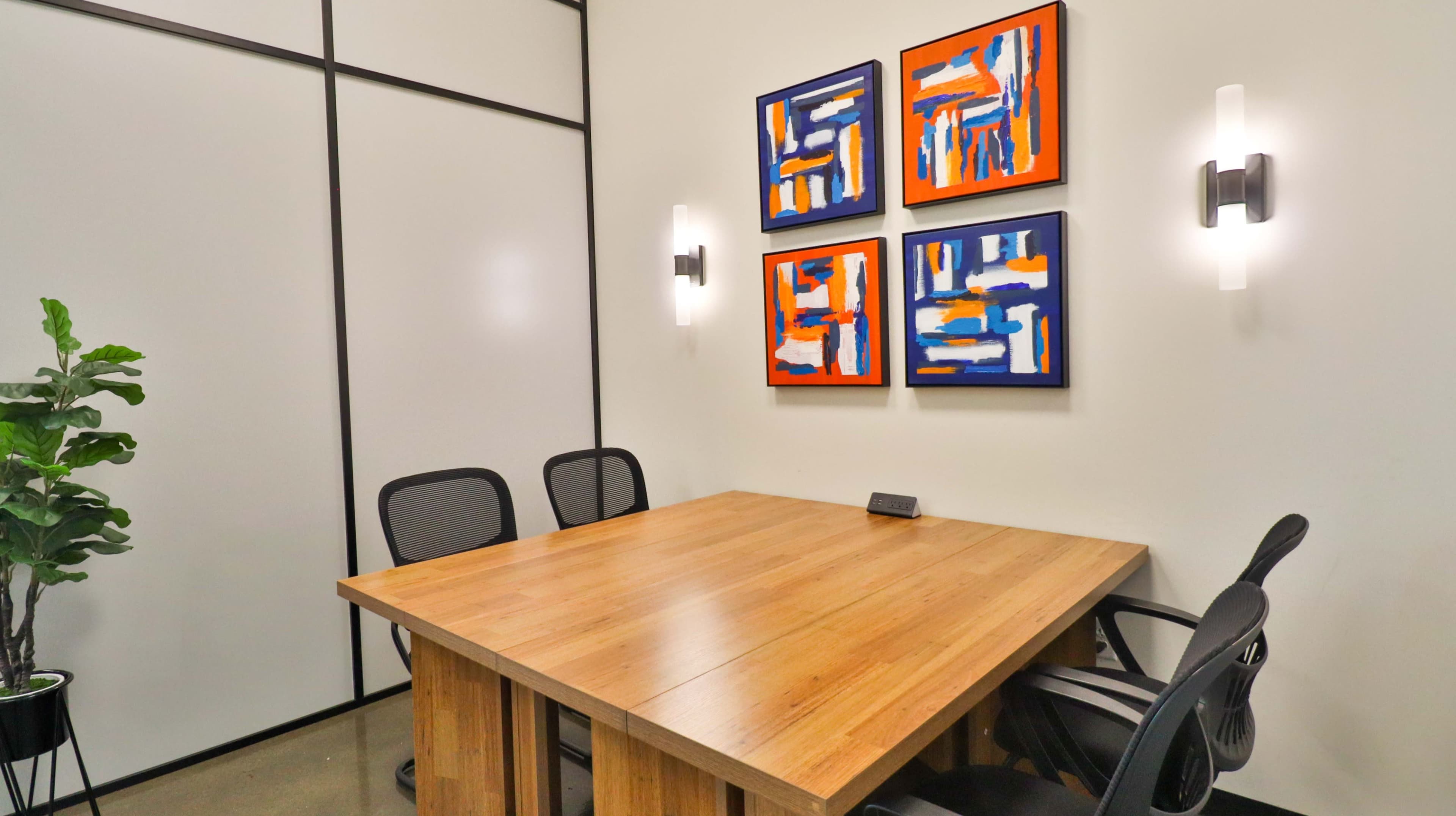 A wooden conference table with four black chairs is positioned in a small meeting room featuring two wall sconces and four colorful abstract paintings.