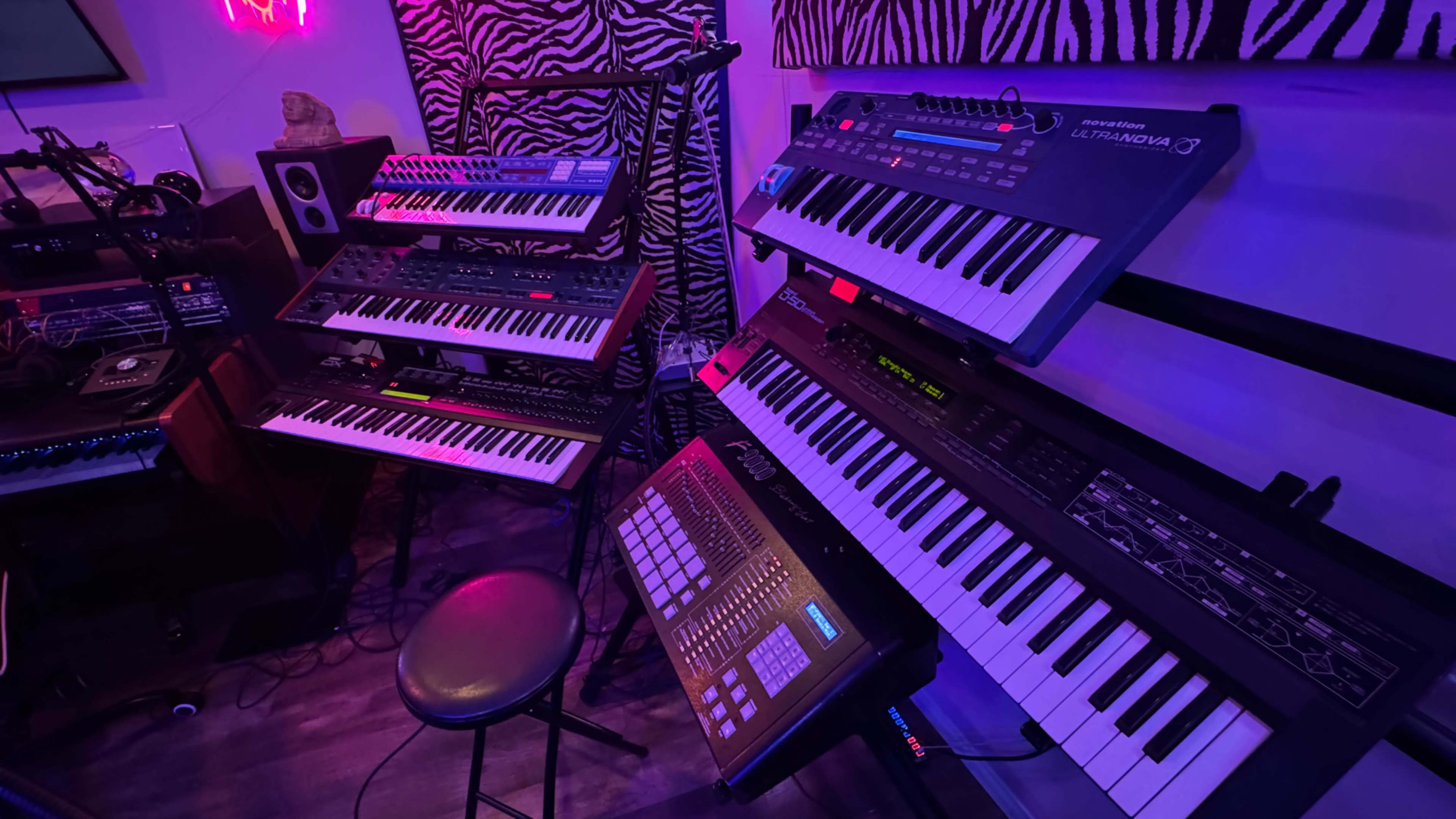 The image shows a collection of synthesizers and a drum machine arranged on stands in a room with a zebra-patterned background.