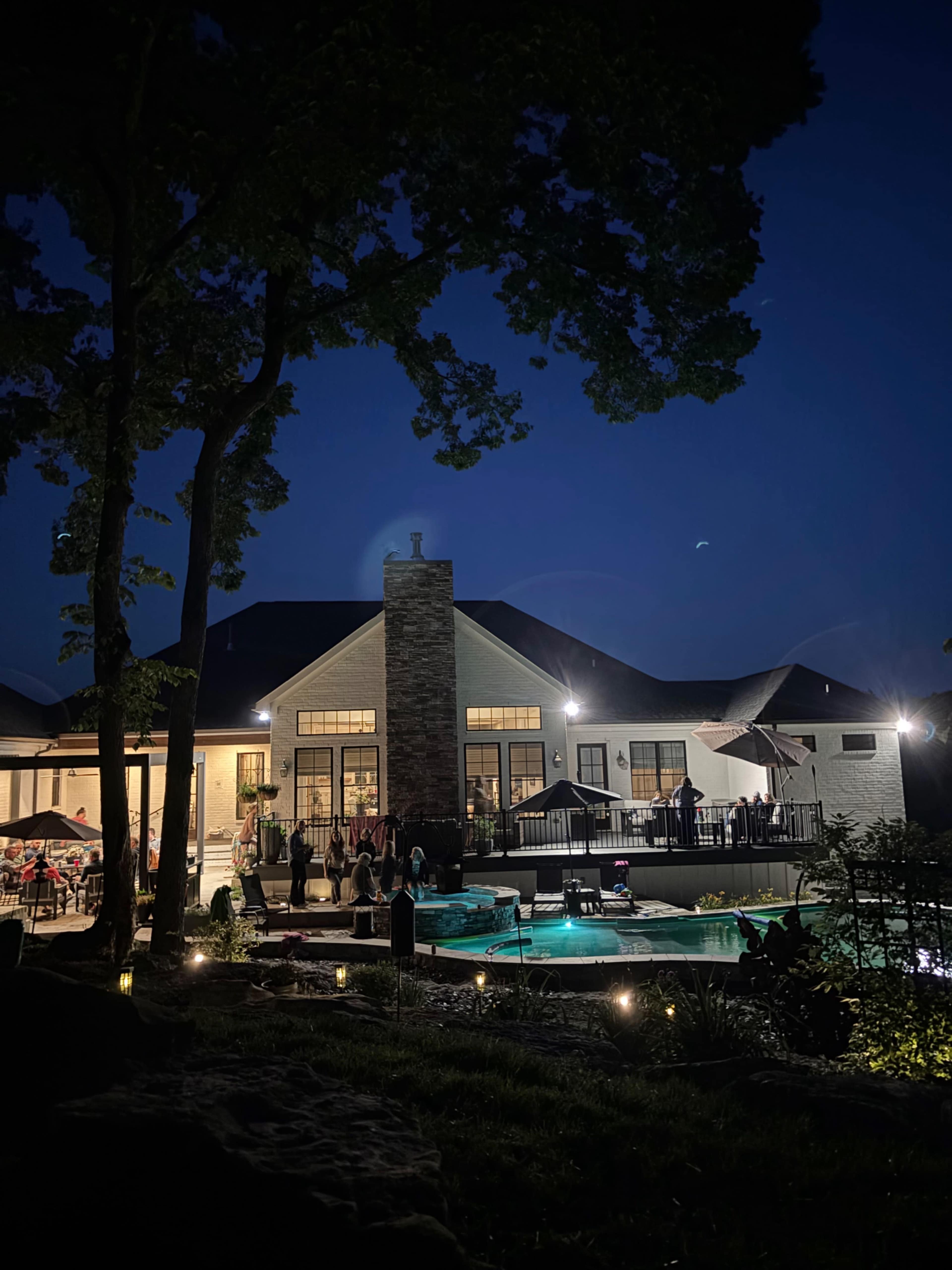 A modern house with a pool and outdoor seating area is illuminated at night, surrounded by trees.
