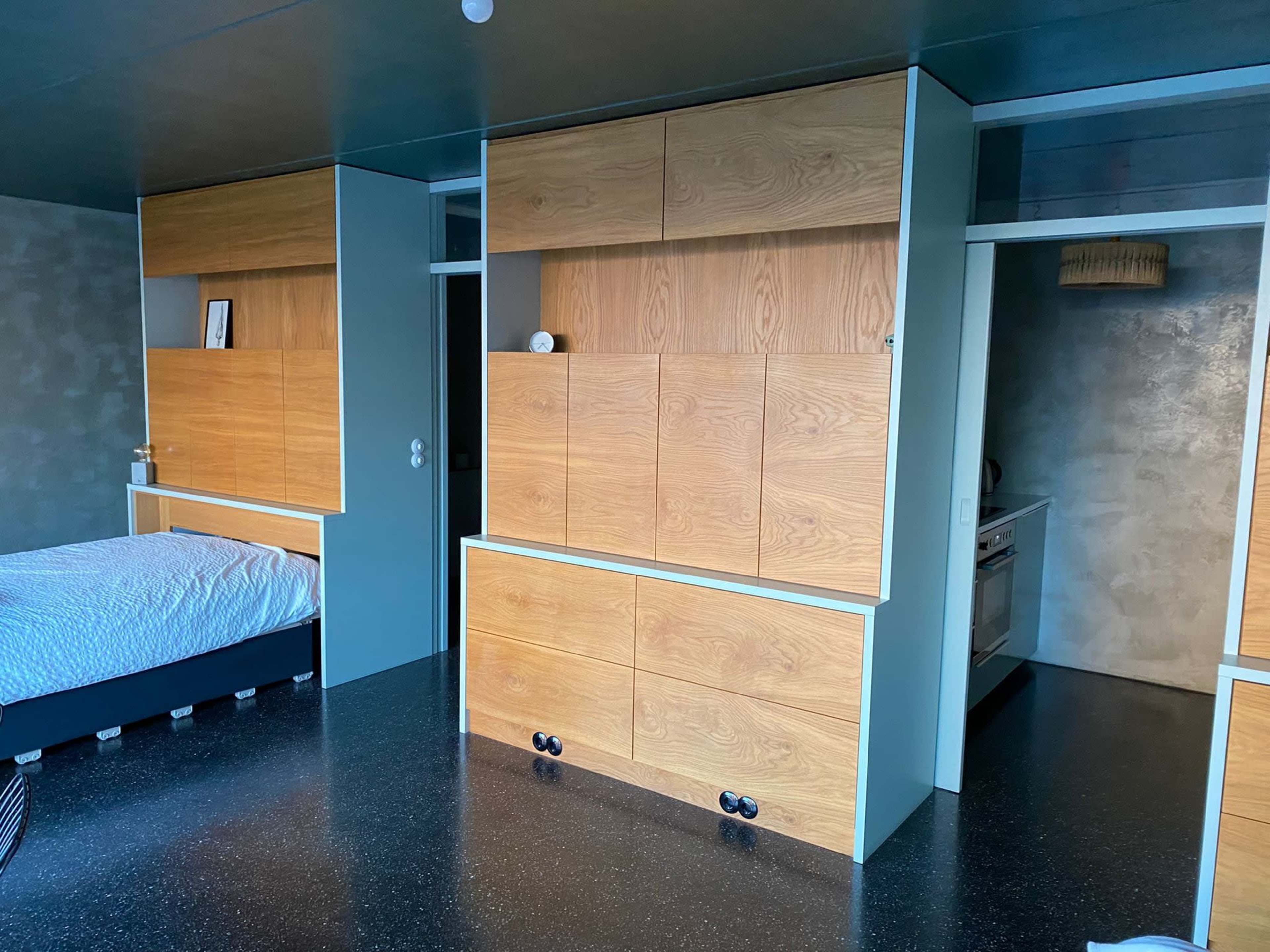 The image shows a modern interior space featuring a bed with a textured white cover, wooden cabinetry, and a central kitchen area in a compact design.