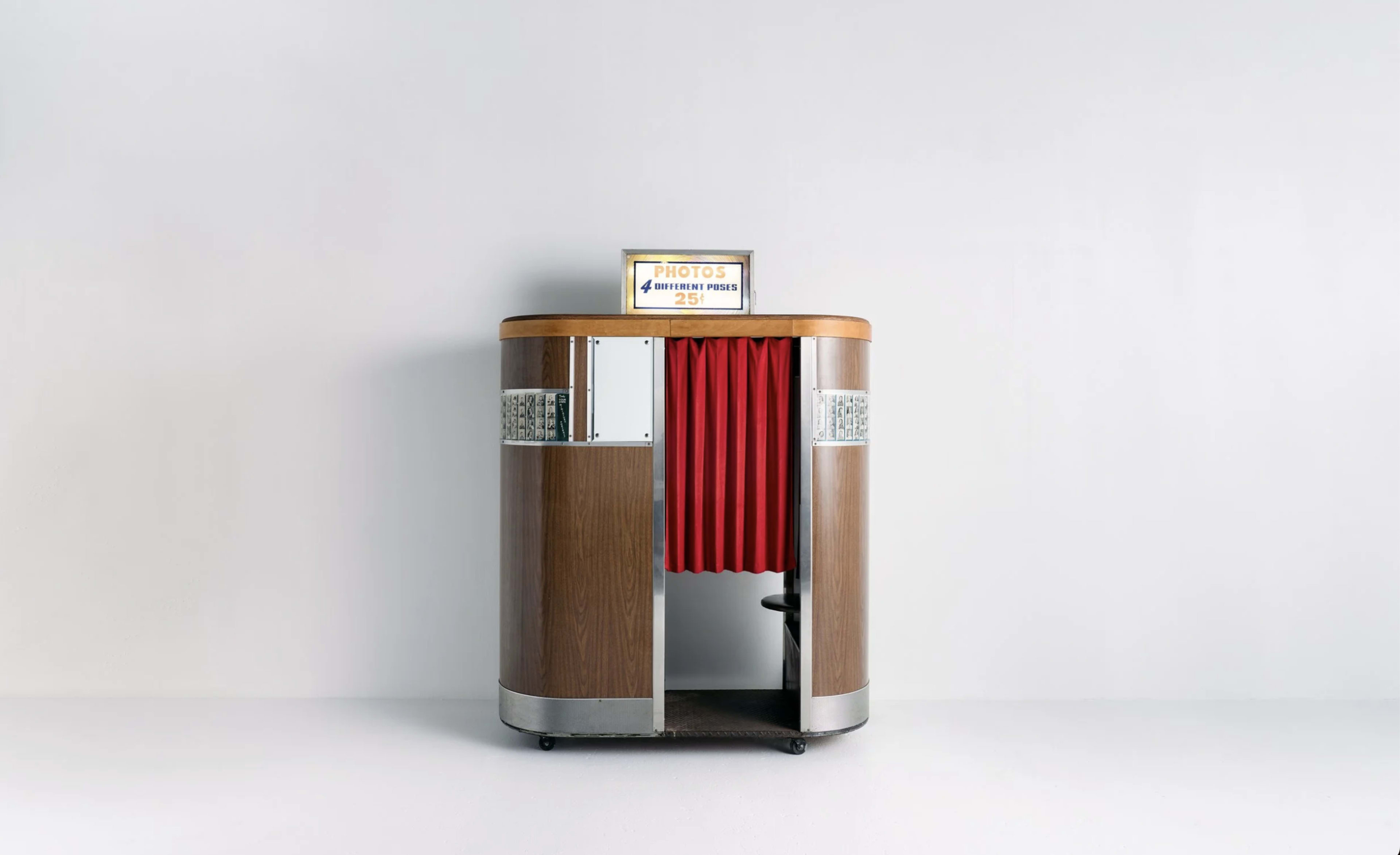 A vintage photo booth with a wooden exterior and red curtains stands against a plain wall.