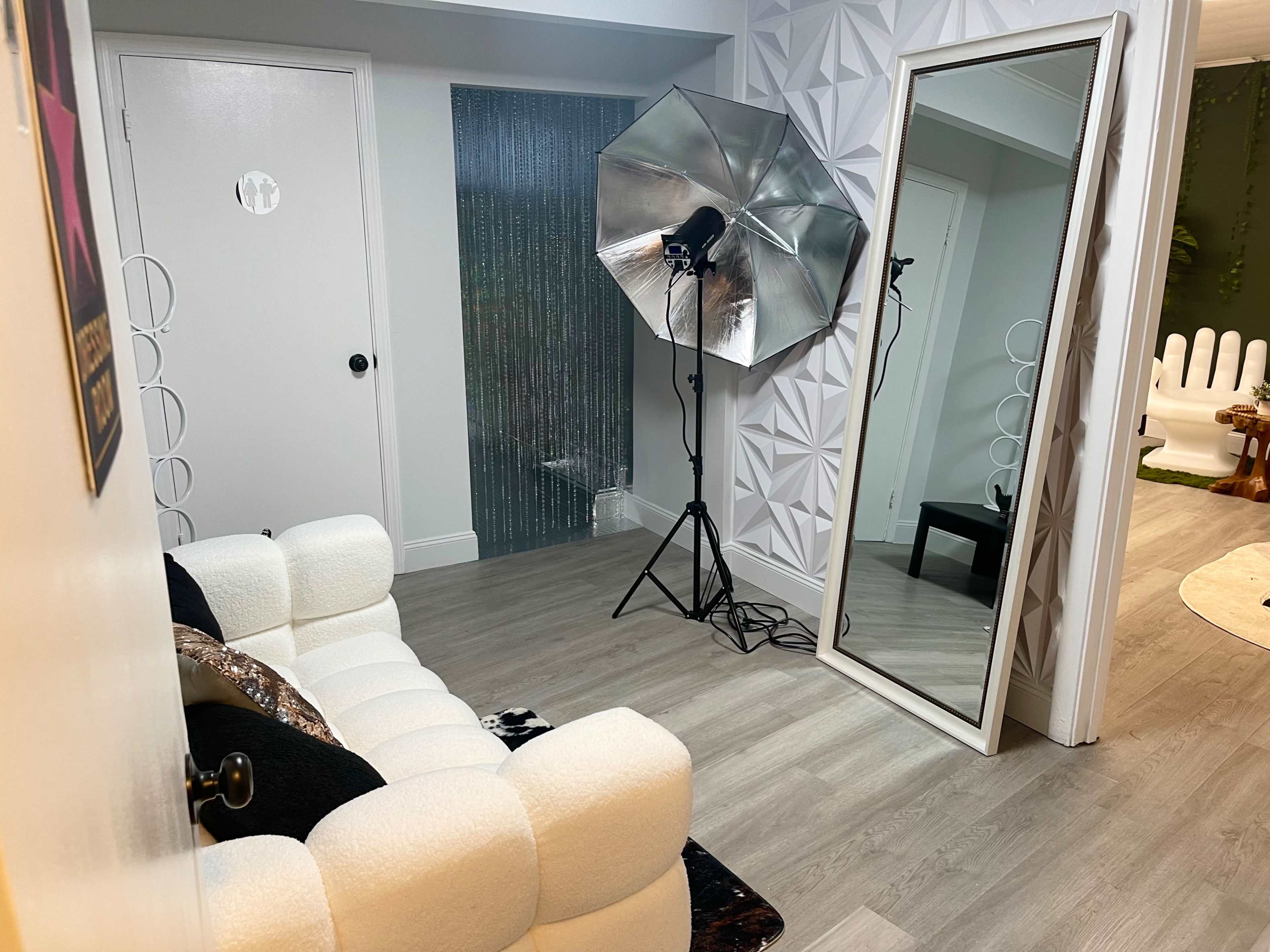A well-lit room featuring a cozy sofa, a large mirror, and a photography umbrella light setup against a wall with a textured design.