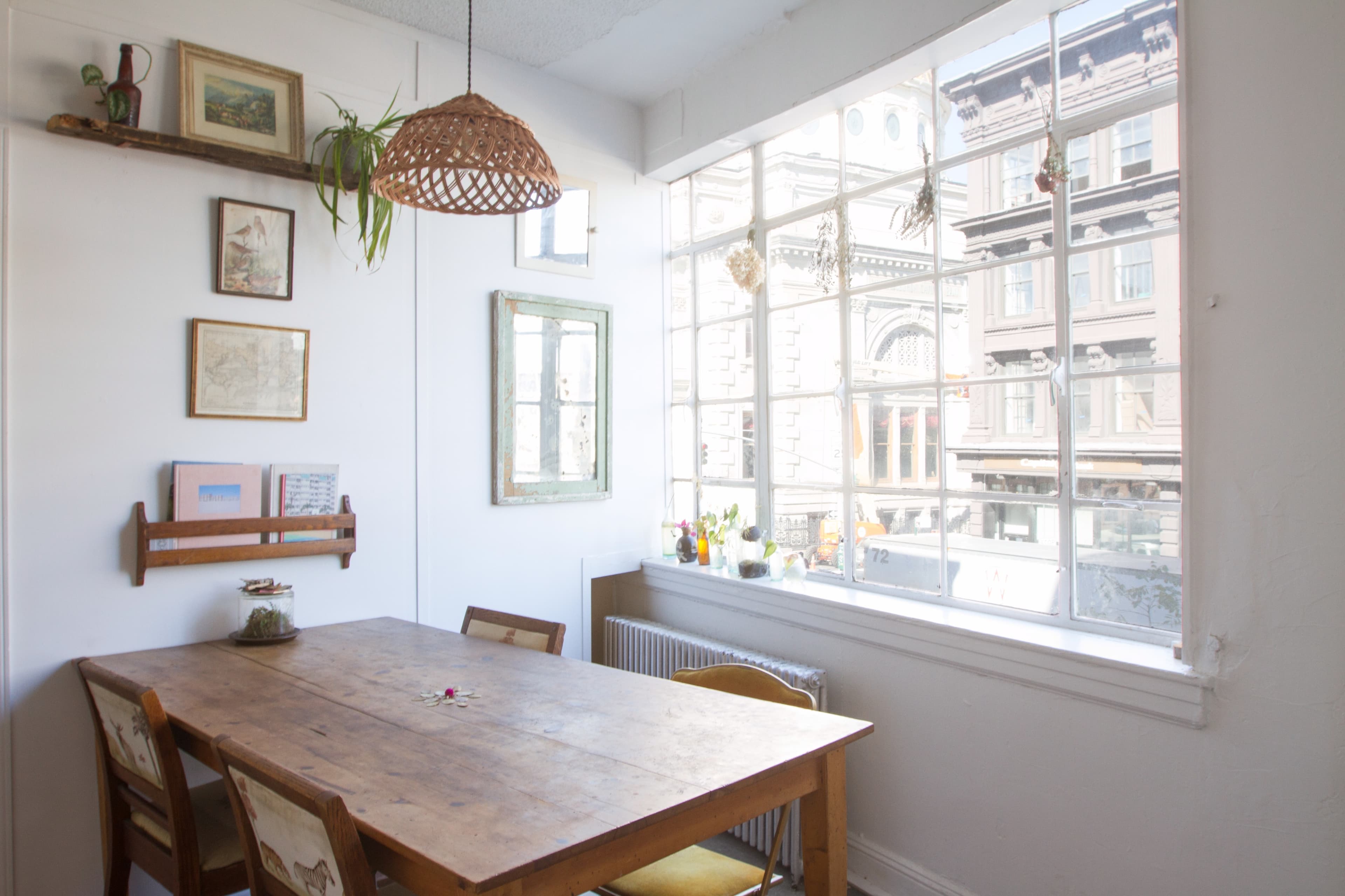 A wooden dining table is set in a bright room with large windows overlooking a city street, showcasing a few wall hangings and plants.