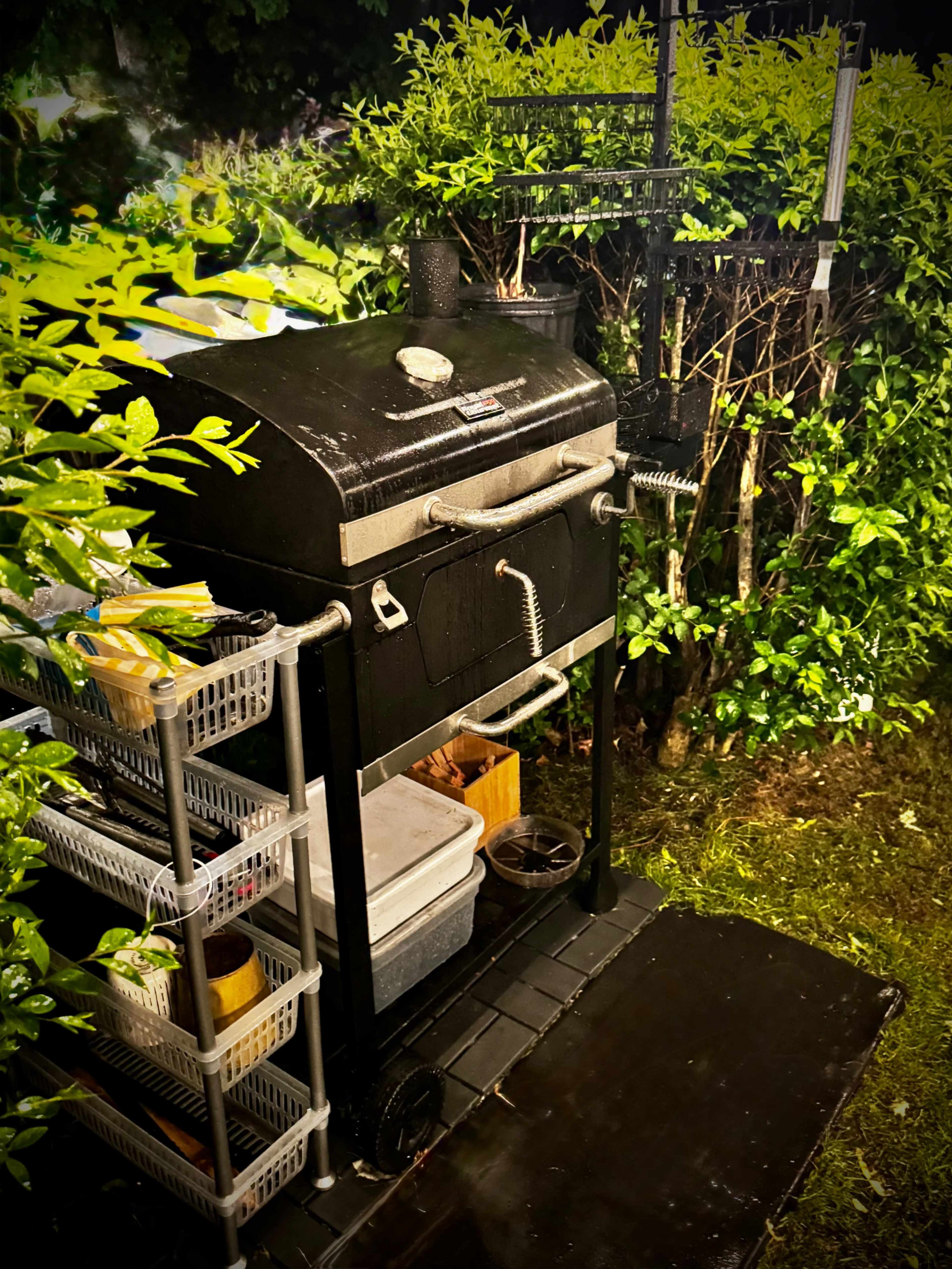 A black grill stands next to a hedge, accompanied by storage shelves filled with cooking supplies.