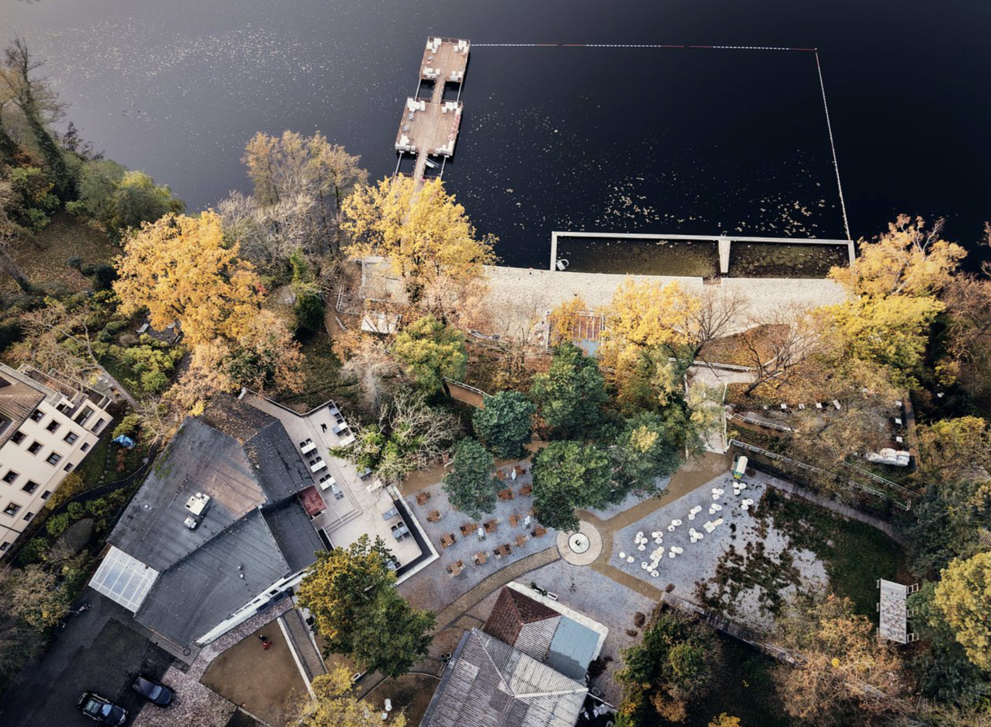 An aerial view shows a lakeside area with wooden docks, fall foliage, and landscaped gardens surrounding a building.