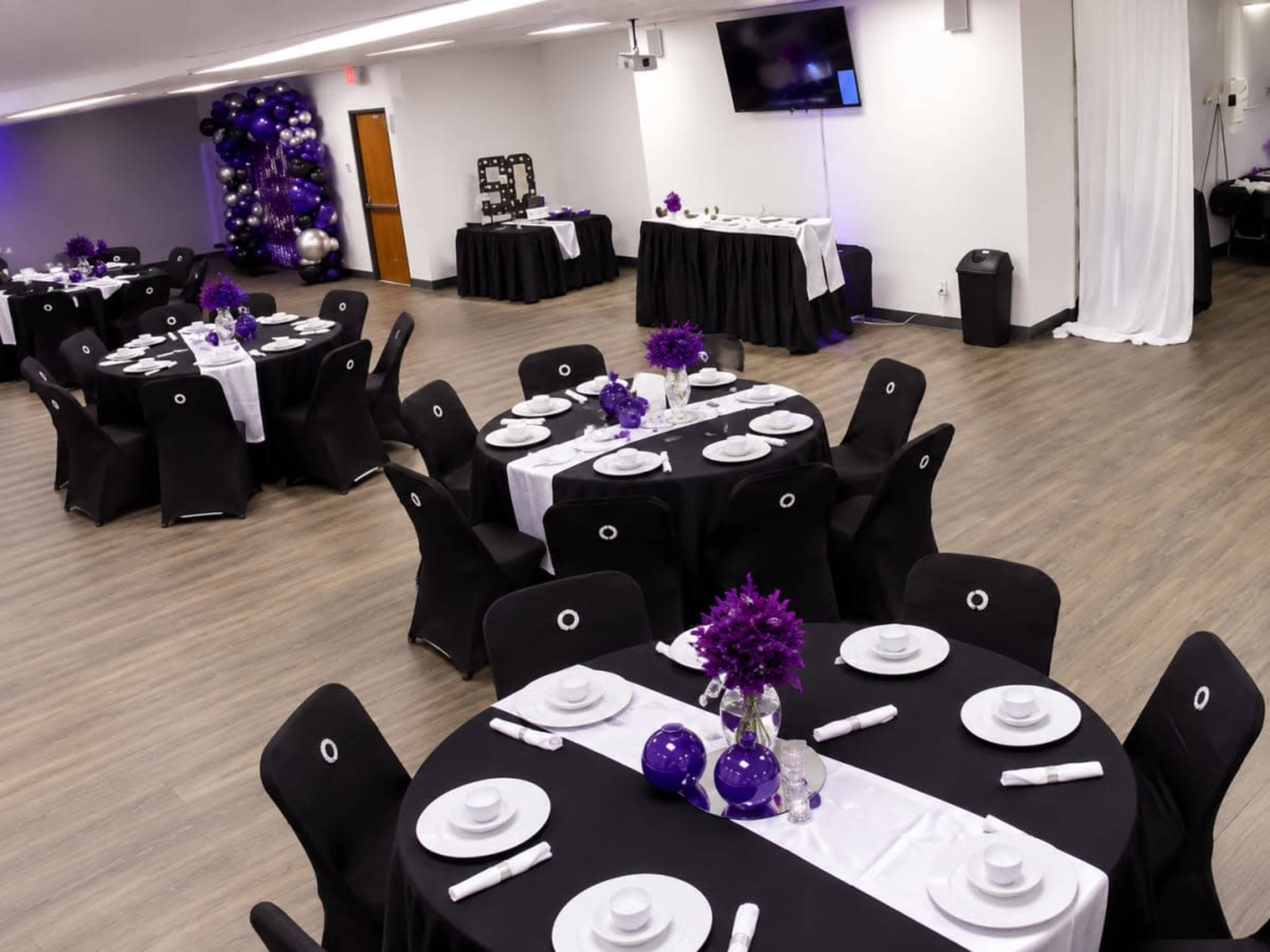 The image depicts a banquet hall set up for an event with round tables covered in black tablecloths, white plates, and purple floral centerpieces.