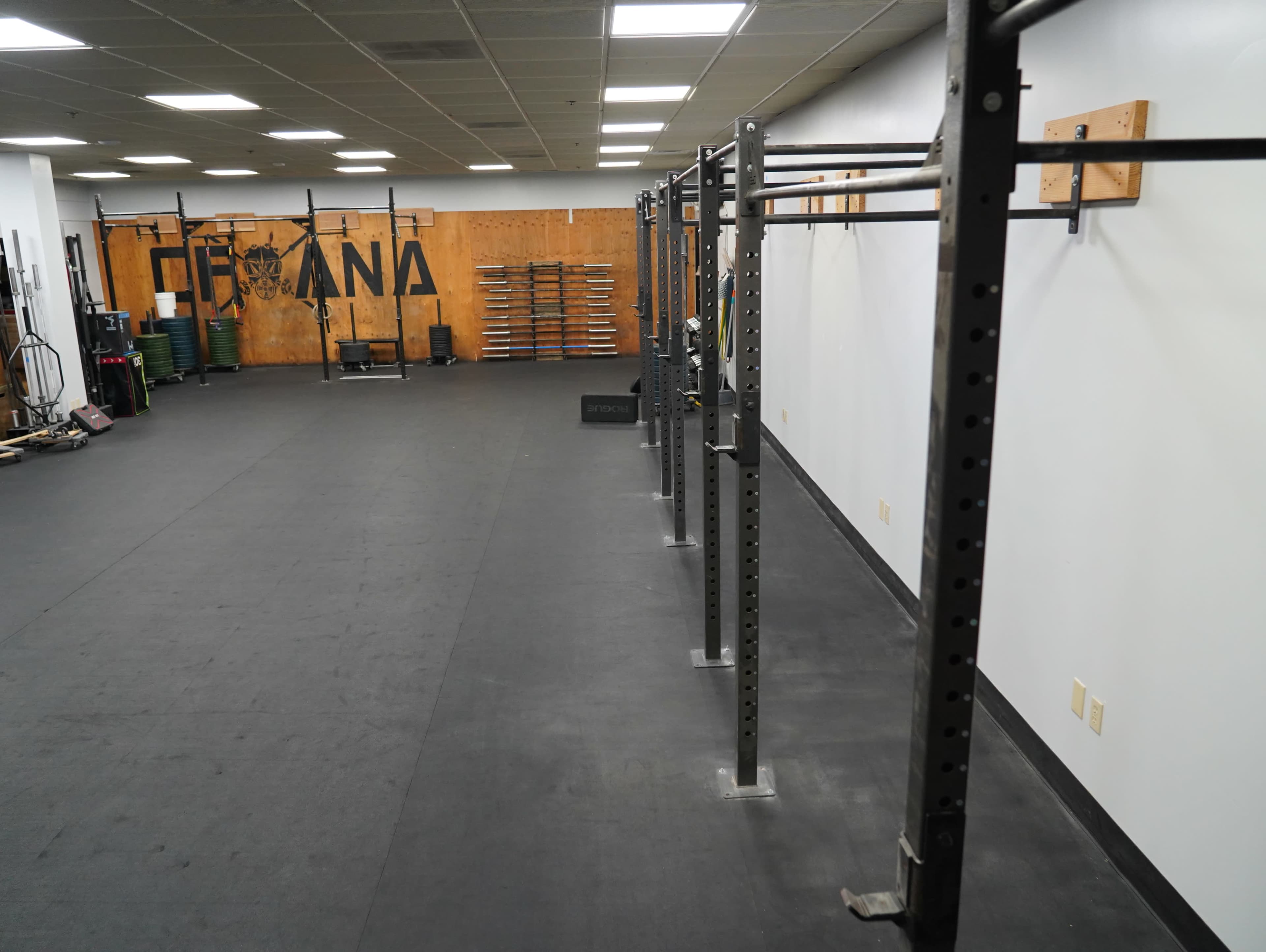 The image shows a spacious gym with pull-up bars, weightlifting equipment, and a rubberized floor, featuring a wooden wall with the word "CROSSFIT" painted on it.