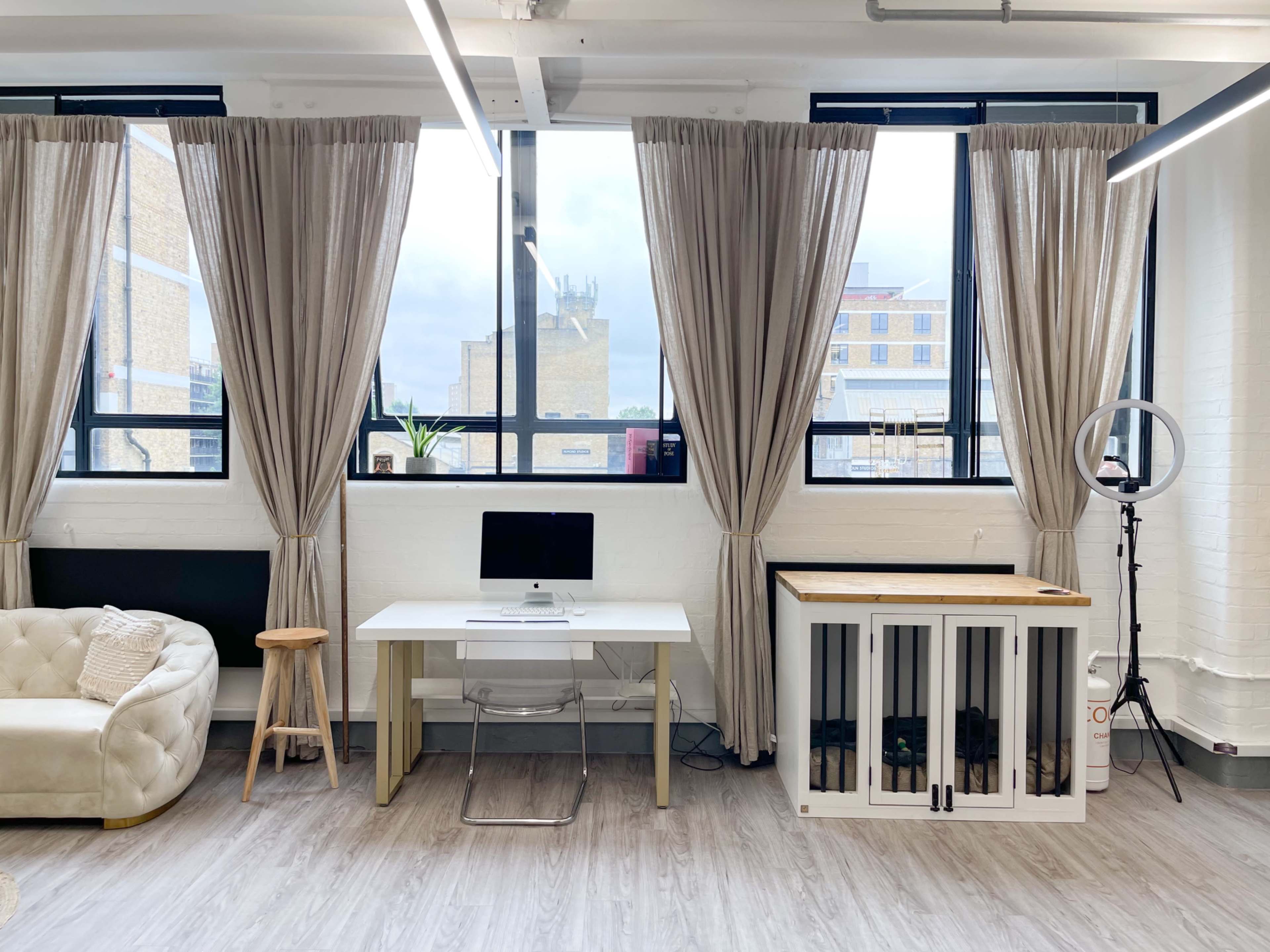 A bright workspace features a desk with a computer, a comfortable sofa, a wooden table, and large windows with draped curtains.