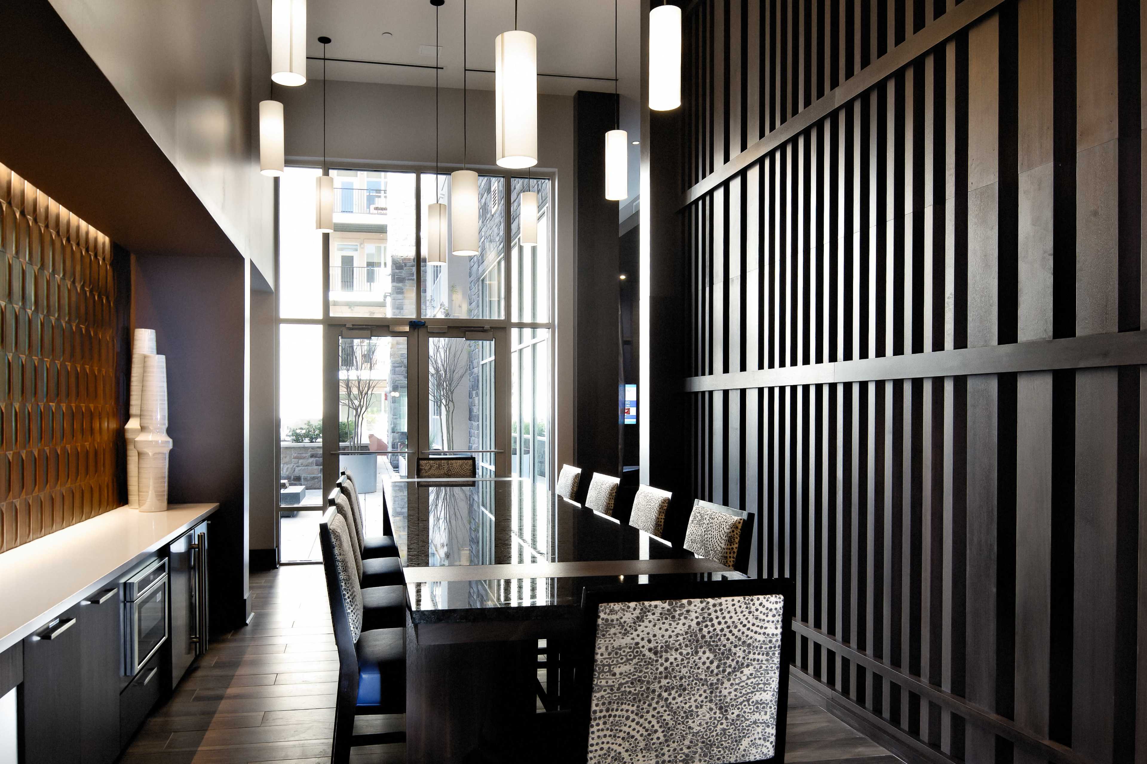 A modern dining area featuring a long dark wood table with patterned chairs, surrounded by tall windows and a textured wooden wall.