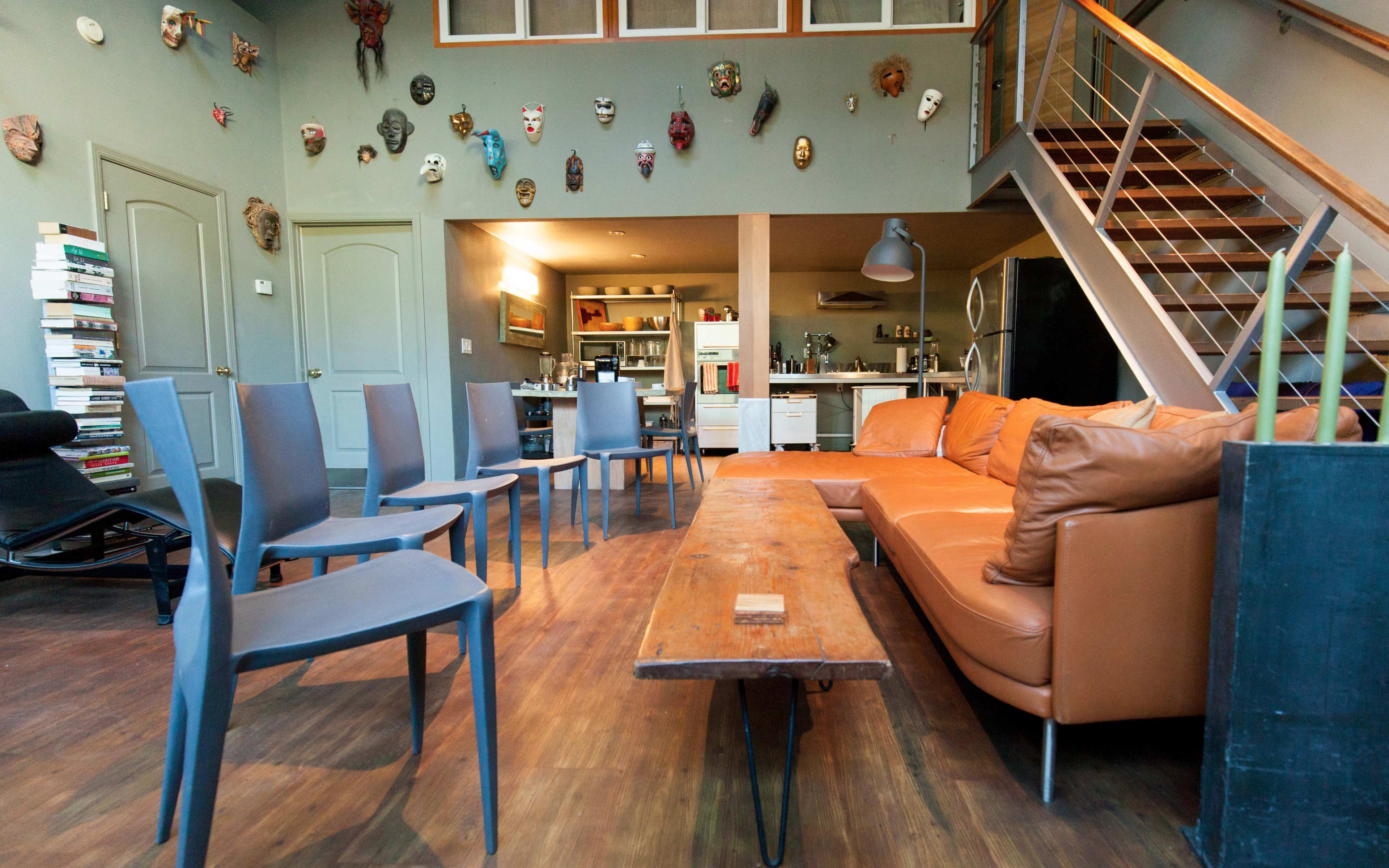 A living room with a brown leather sofa, a wooden coffee table, several gray chairs arranged around the room, and a staircase leading to an upper level adorned with various masks on the walls.
