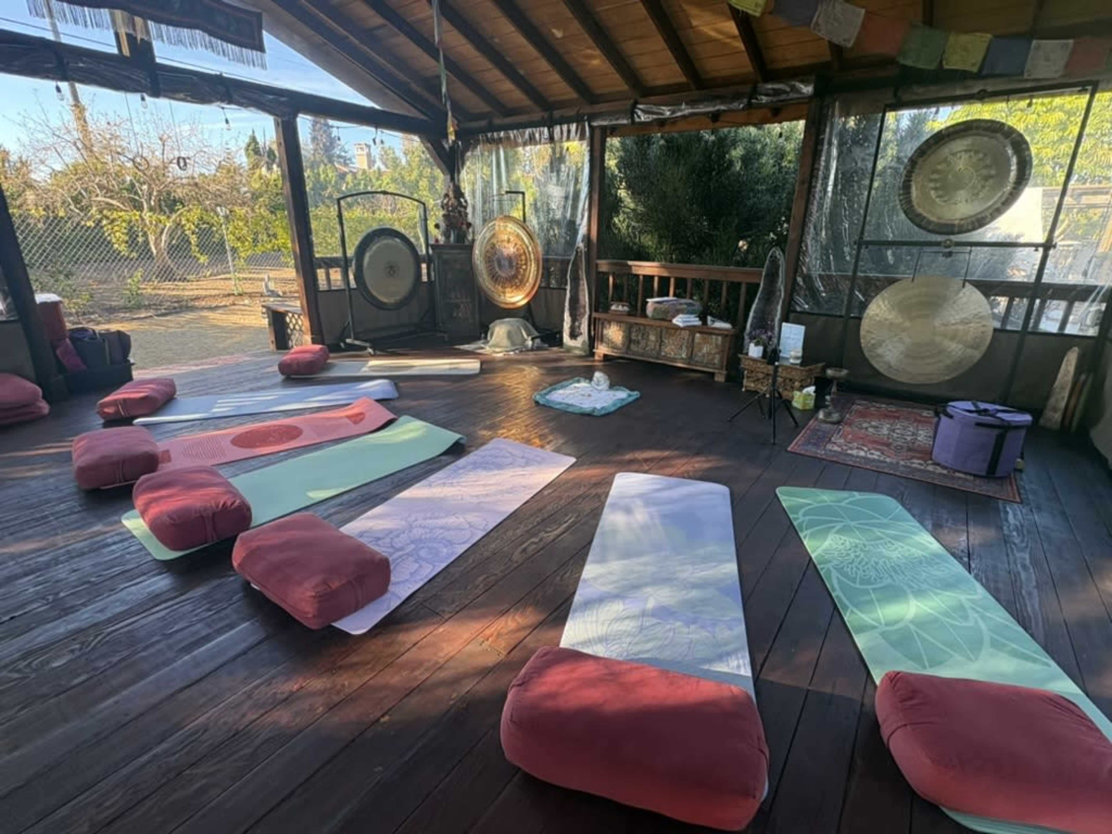 The image shows a wooden deck area arranged with mats and cushions for a wellness session, surrounded by various large instruments and greenery.