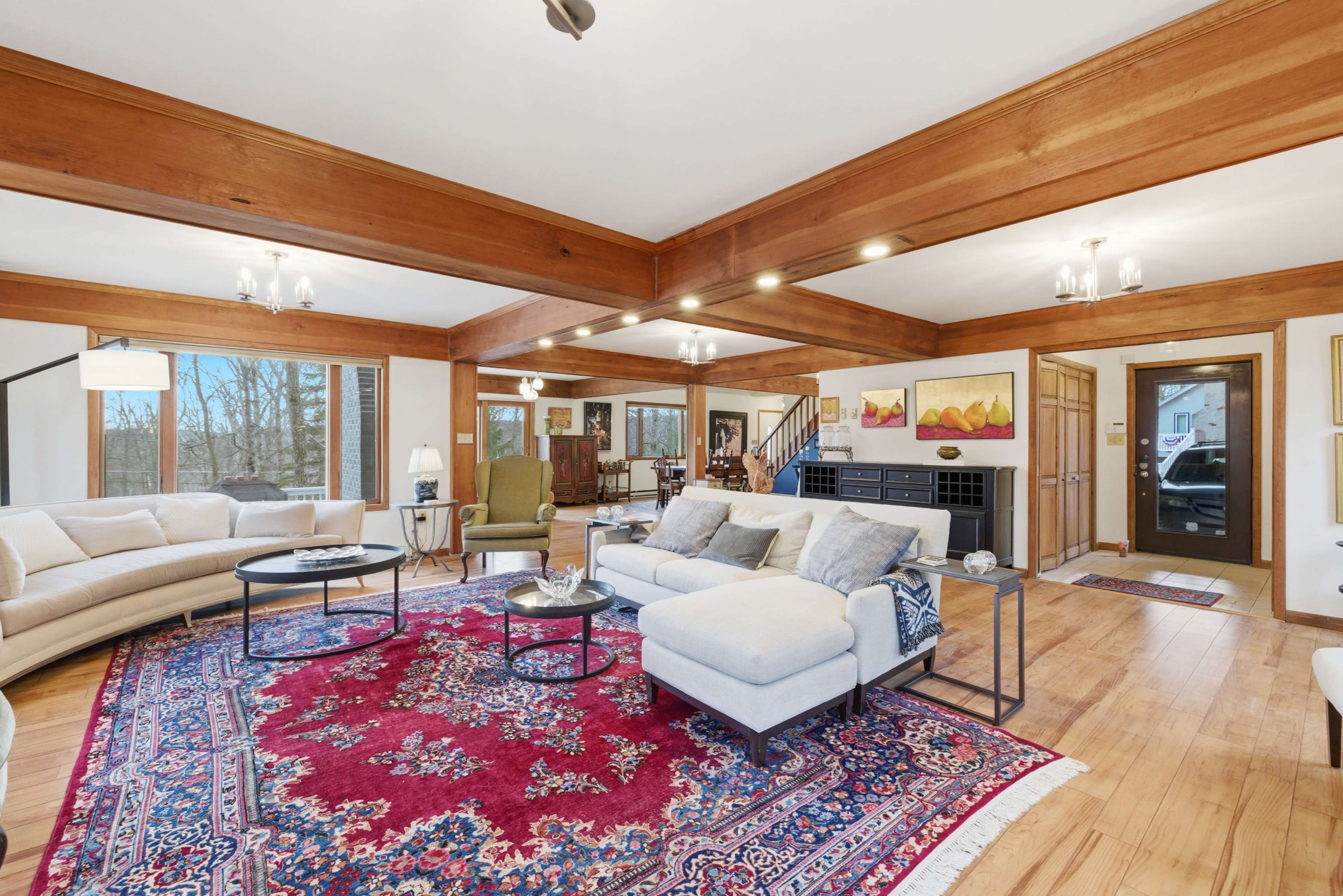 The spacious living room features wooden beams, large windows, and a decorative area rug, furnished with a sectional sofa and a coffee table.