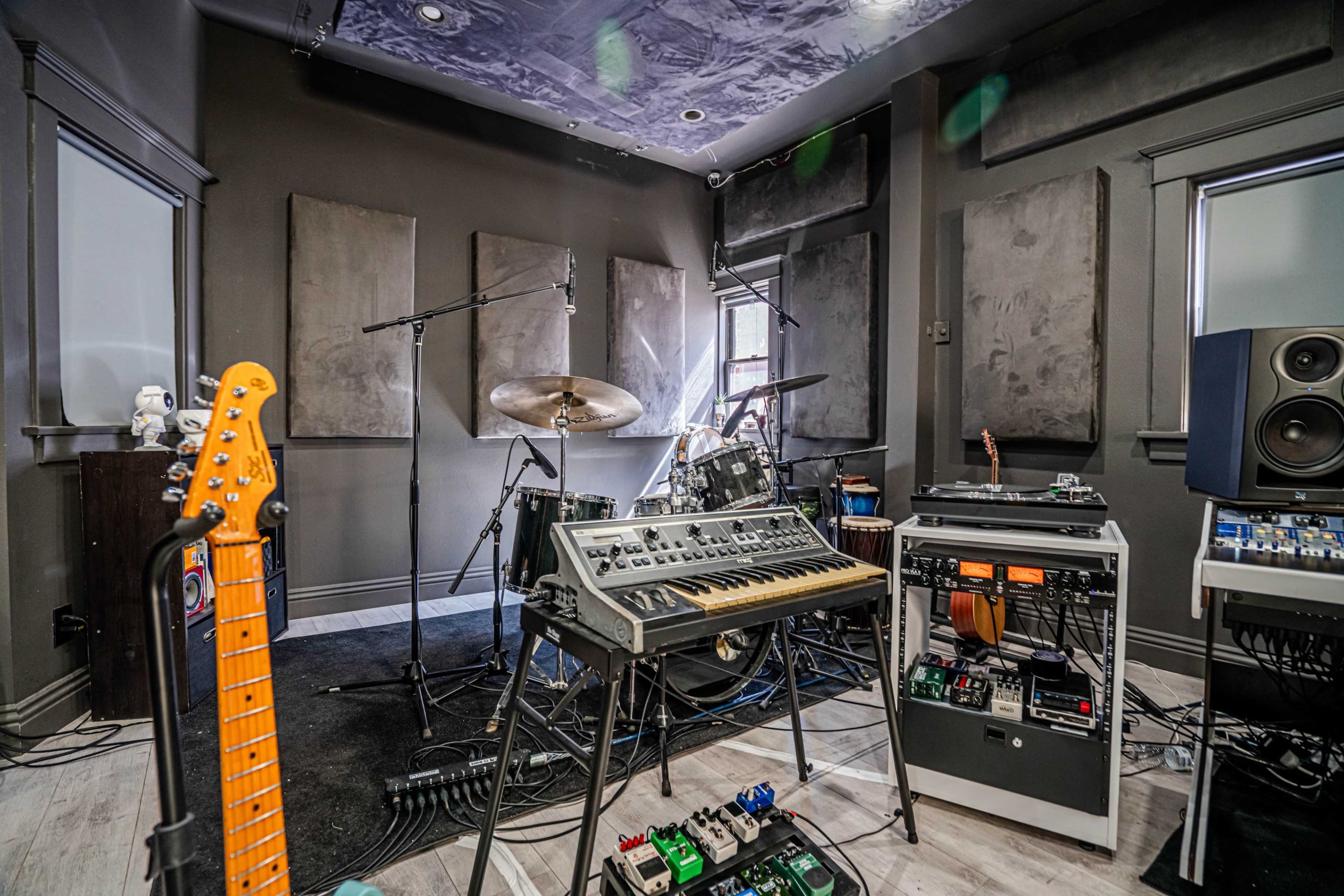 The image shows a music studio filled with various instruments, including a guitar, a synthesizer, and a drum set, surrounded by soundproofing panels on the walls.