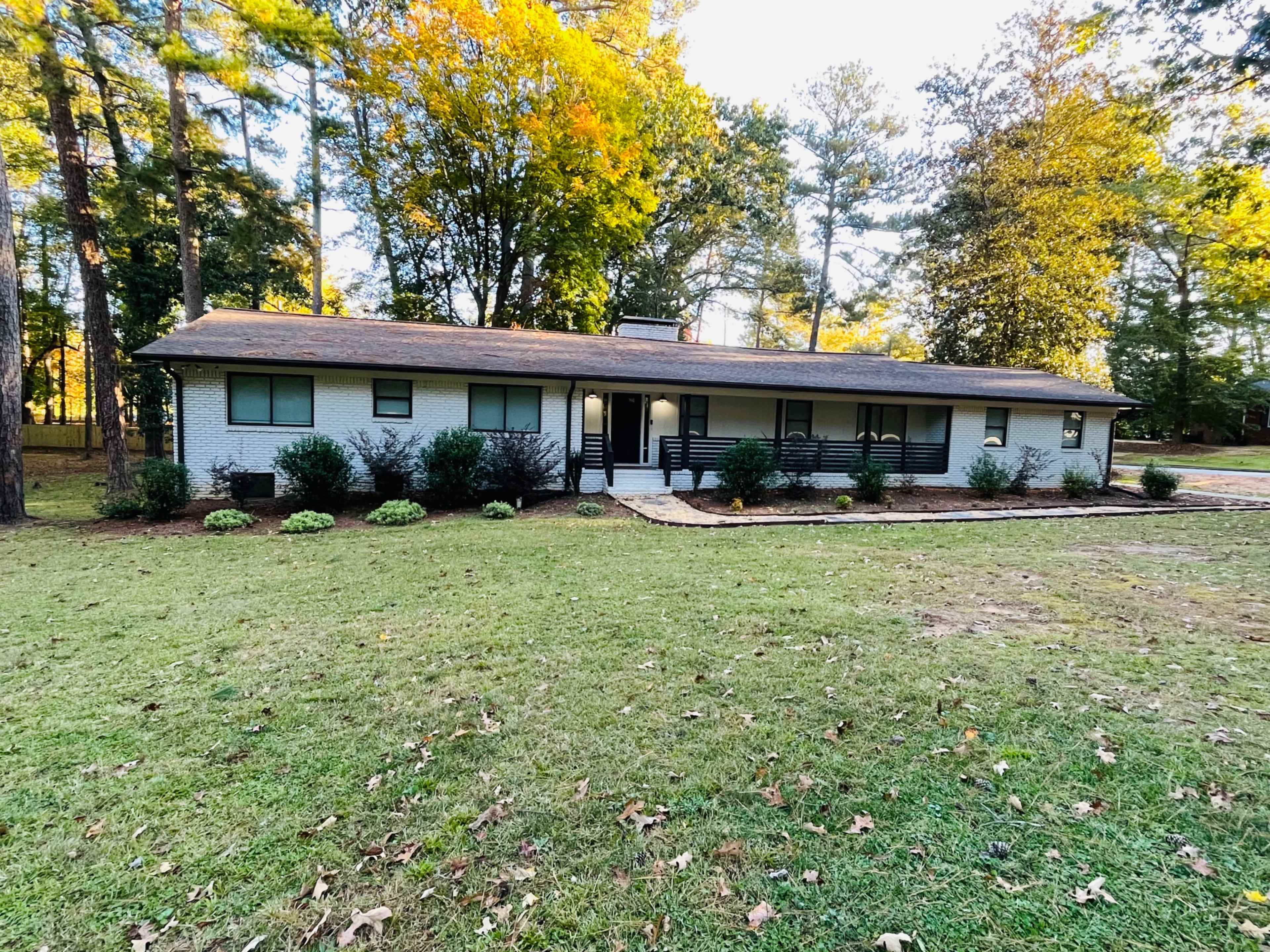 A single-story, mid-century modern house with a flat roof is surrounded by greenery and trees in a residential setting.