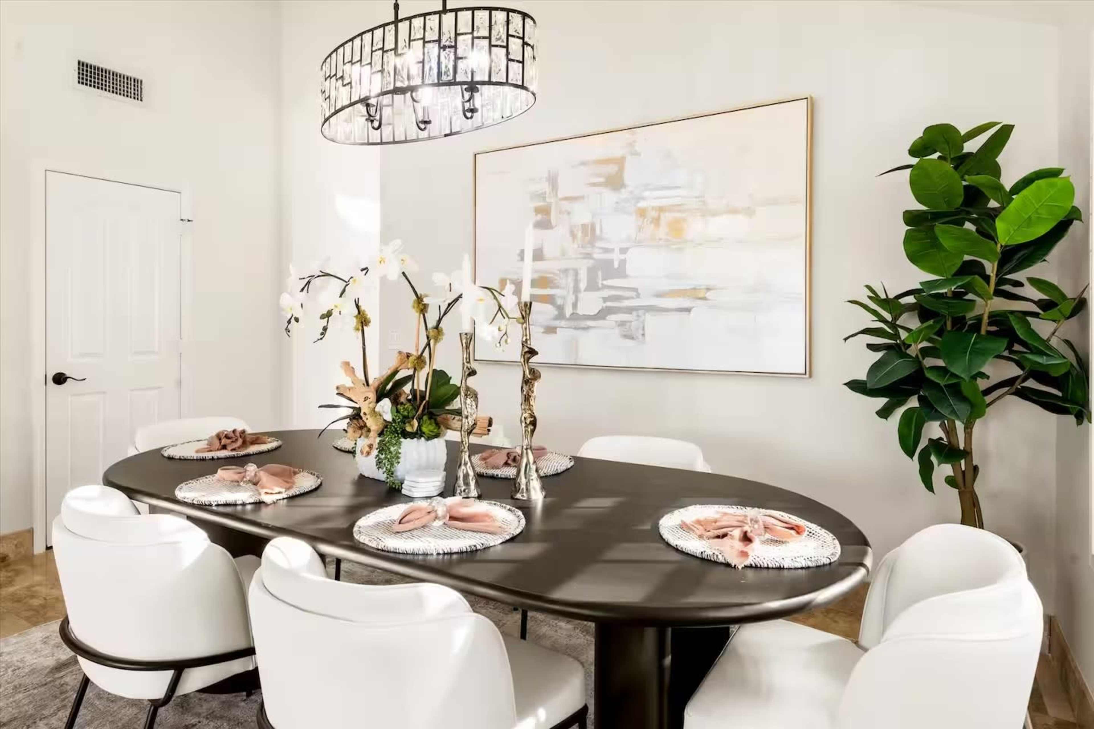 A modern dining room features a large oval table set with plates and centerpieces, surrounded by white chairs and illuminated by a decorative chandelier.