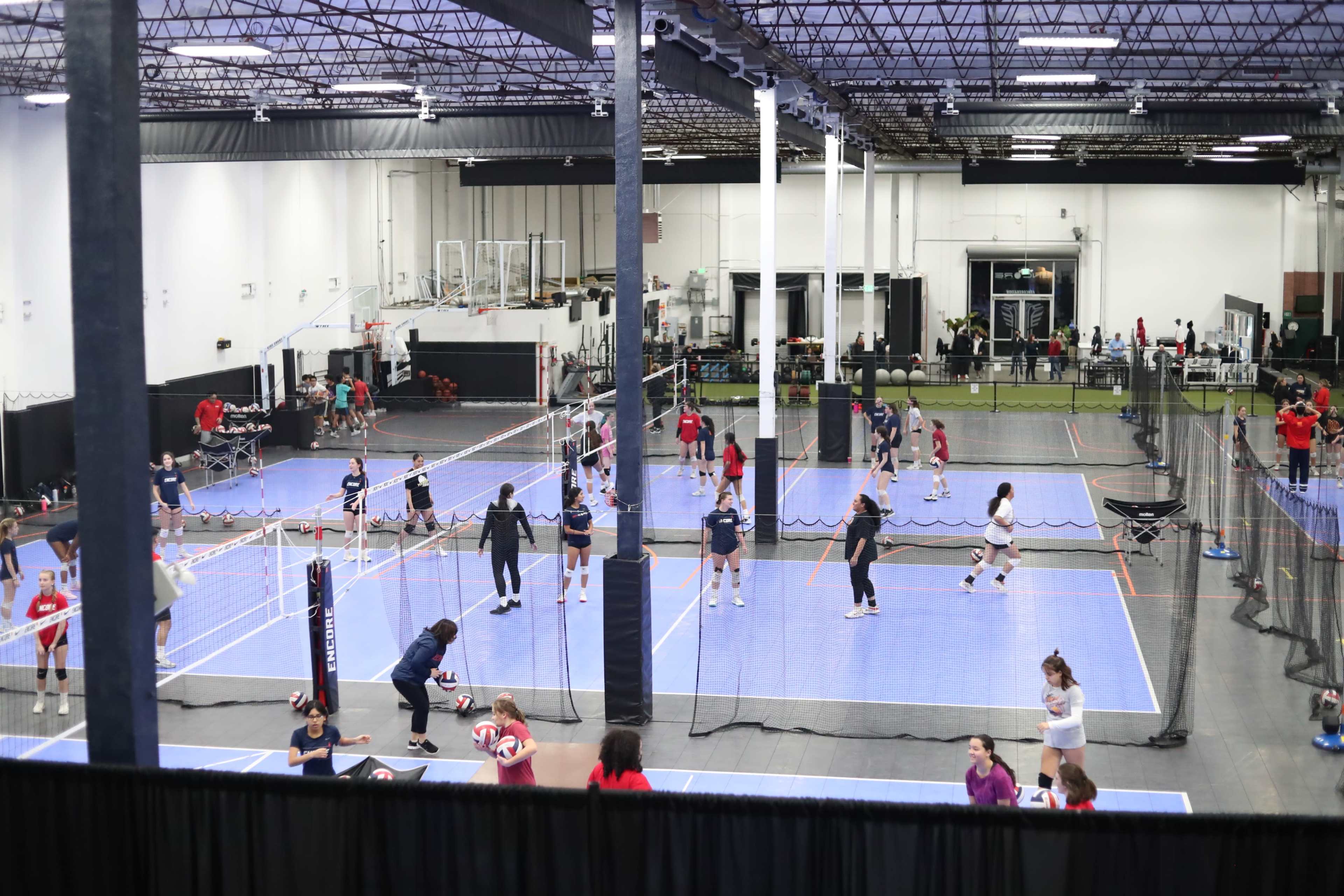 A large indoor gymnasium features multiple volleyball courts where players are actively participating in drills and matches.