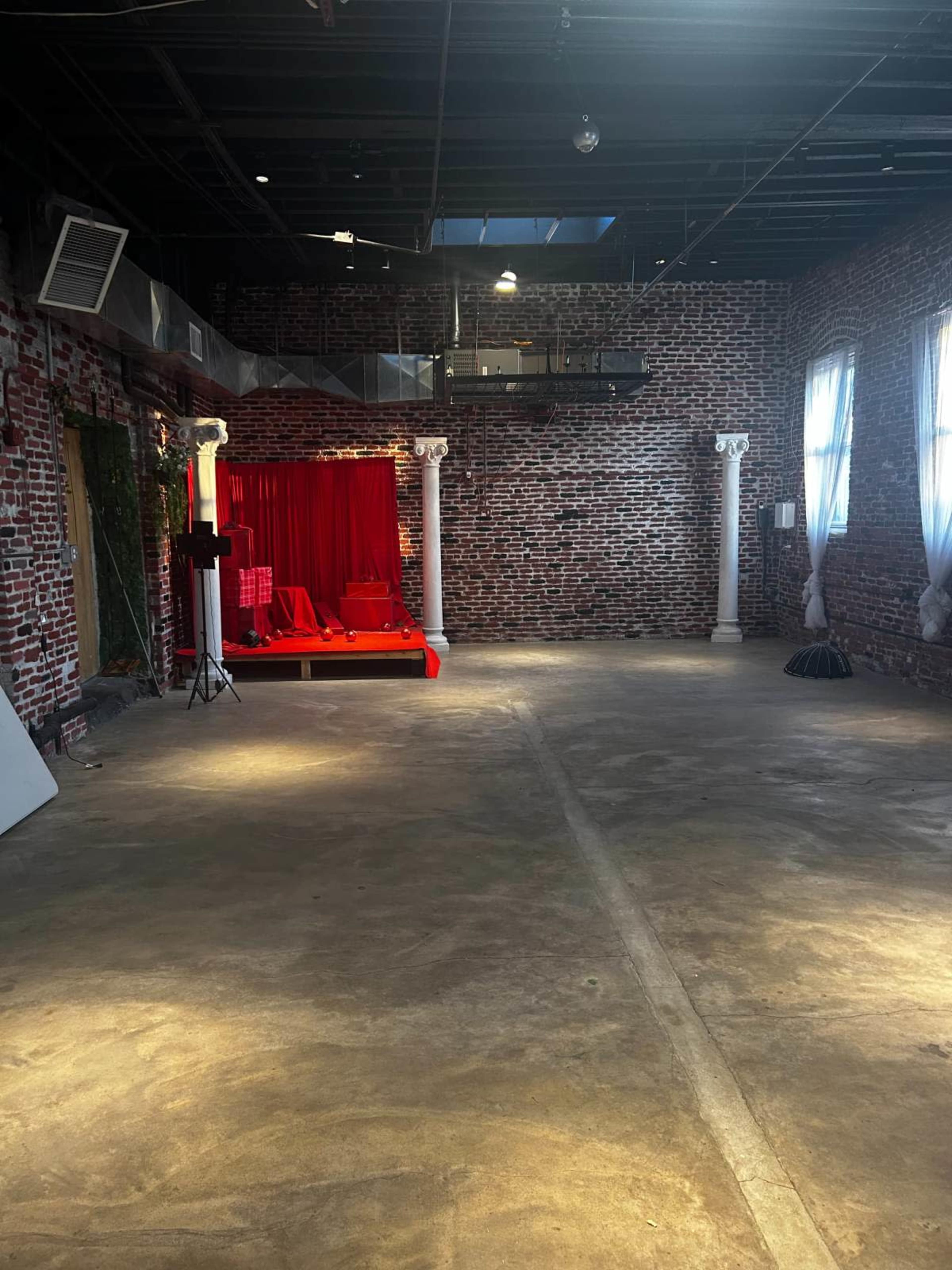 The image shows an empty industrial space with exposed brick walls, a concrete floor, and a red draped area featuring a seating arrangement.