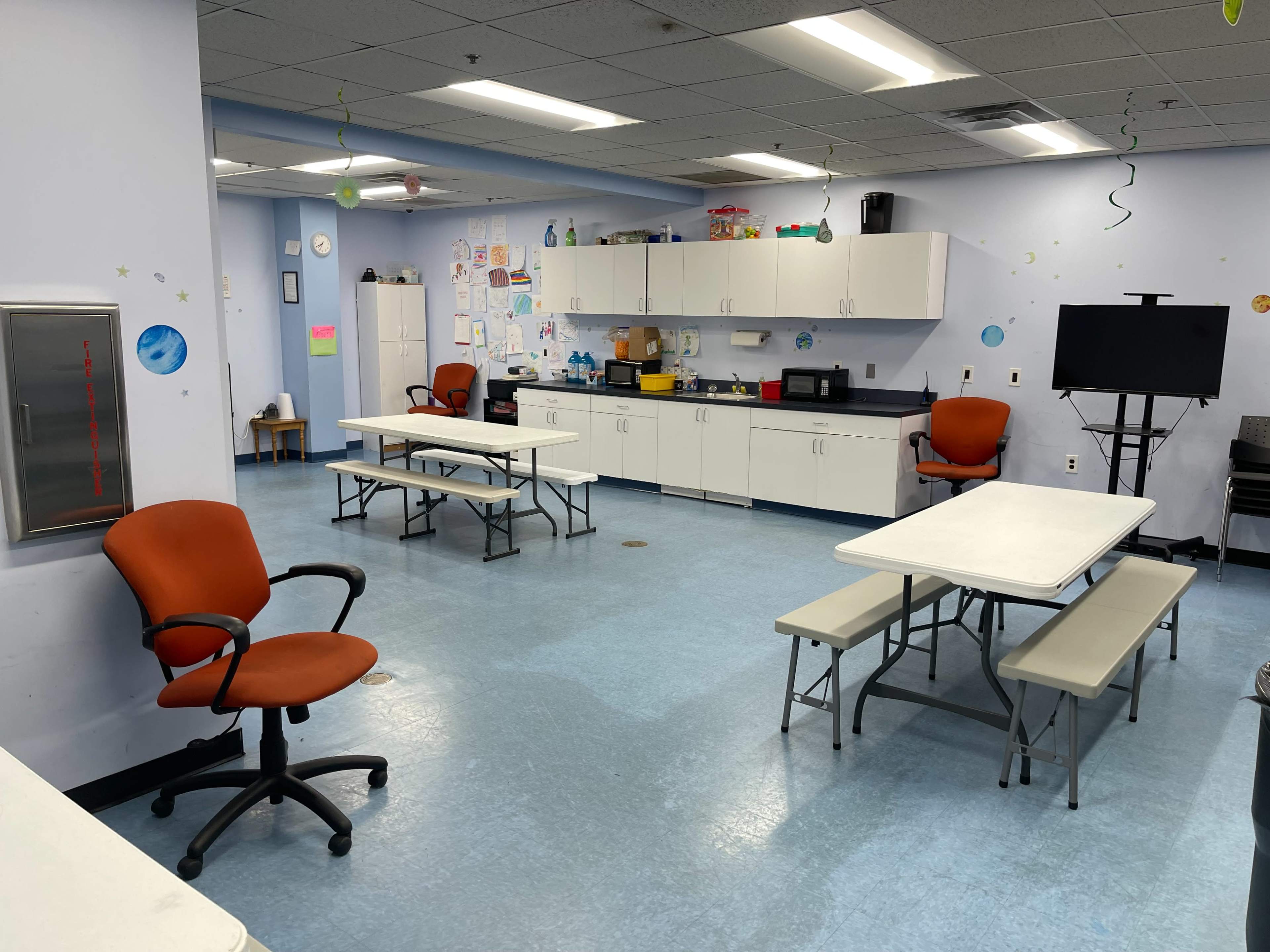 A bright room with several tables and chairs, a kitchenette area, and decorations on the walls.