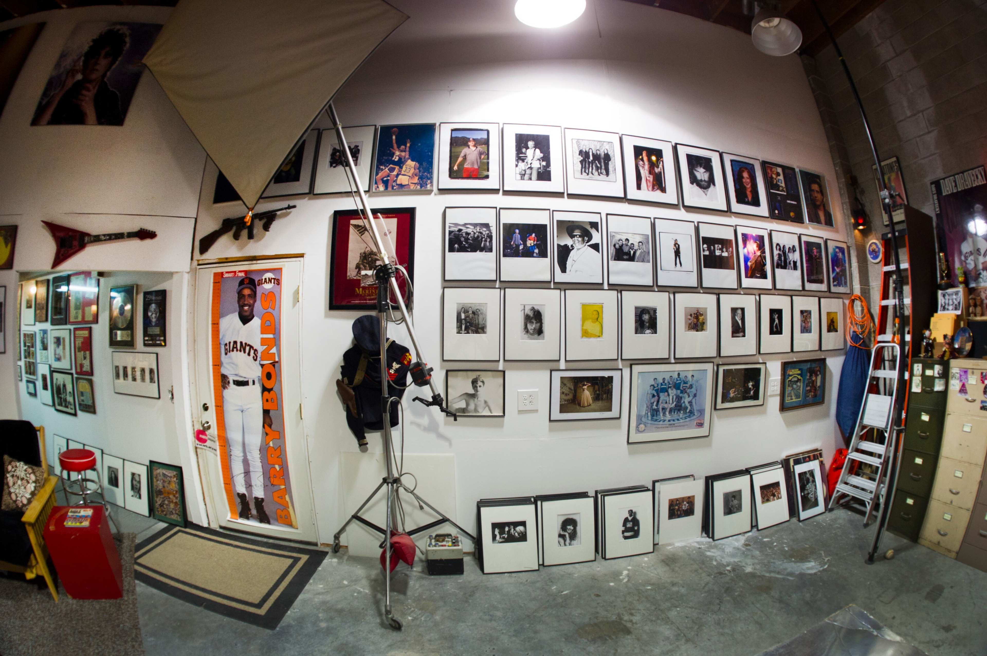 A wall covered with framed photographs and artwork, featuring various celebrity portraits and sports memorabilia, alongside a photography backdrop and equipment.