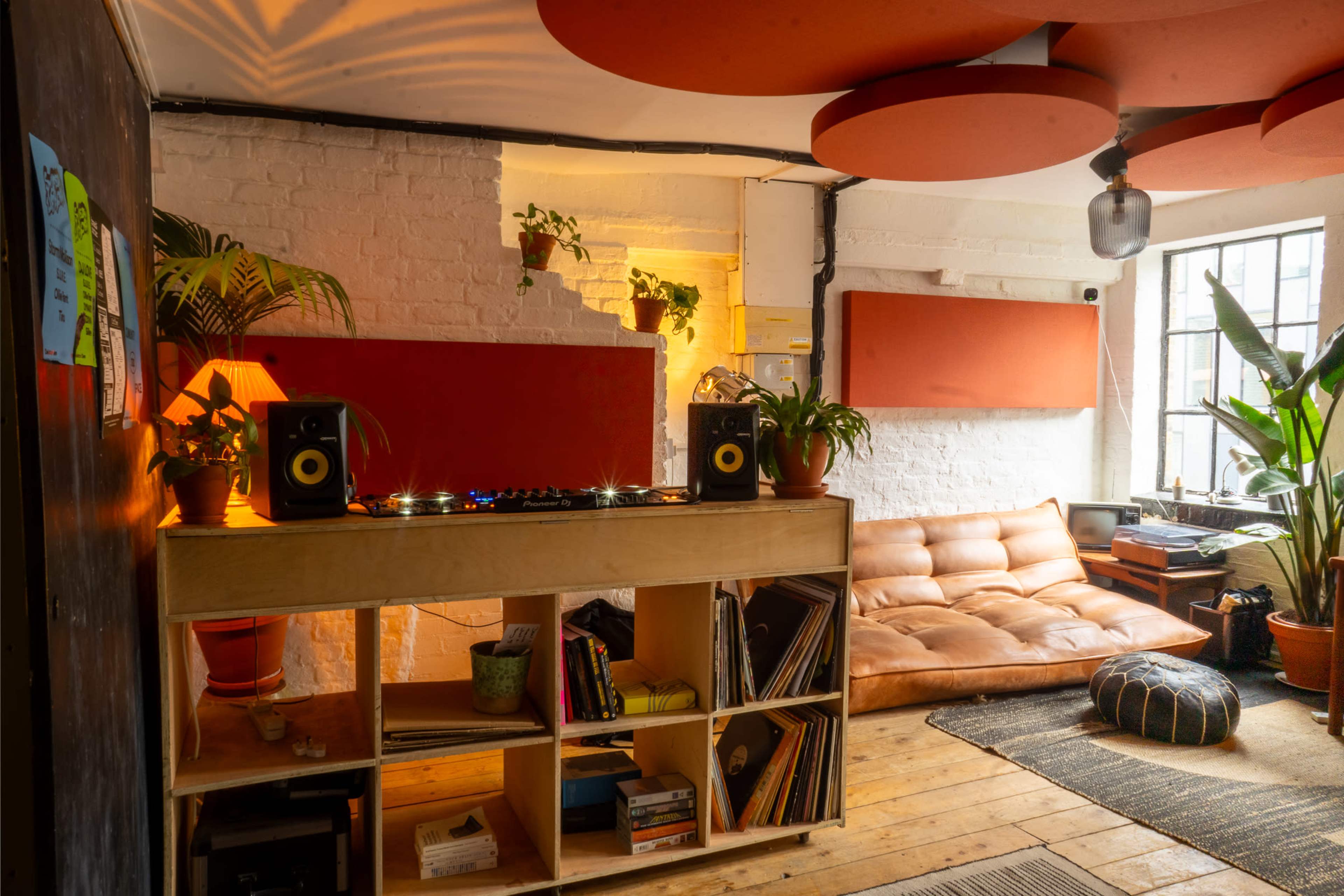 The image shows a cozy interior of a room featuring a wooden shelf with vinyl records, a DJ setup, a brown sofa, and various indoor plants.