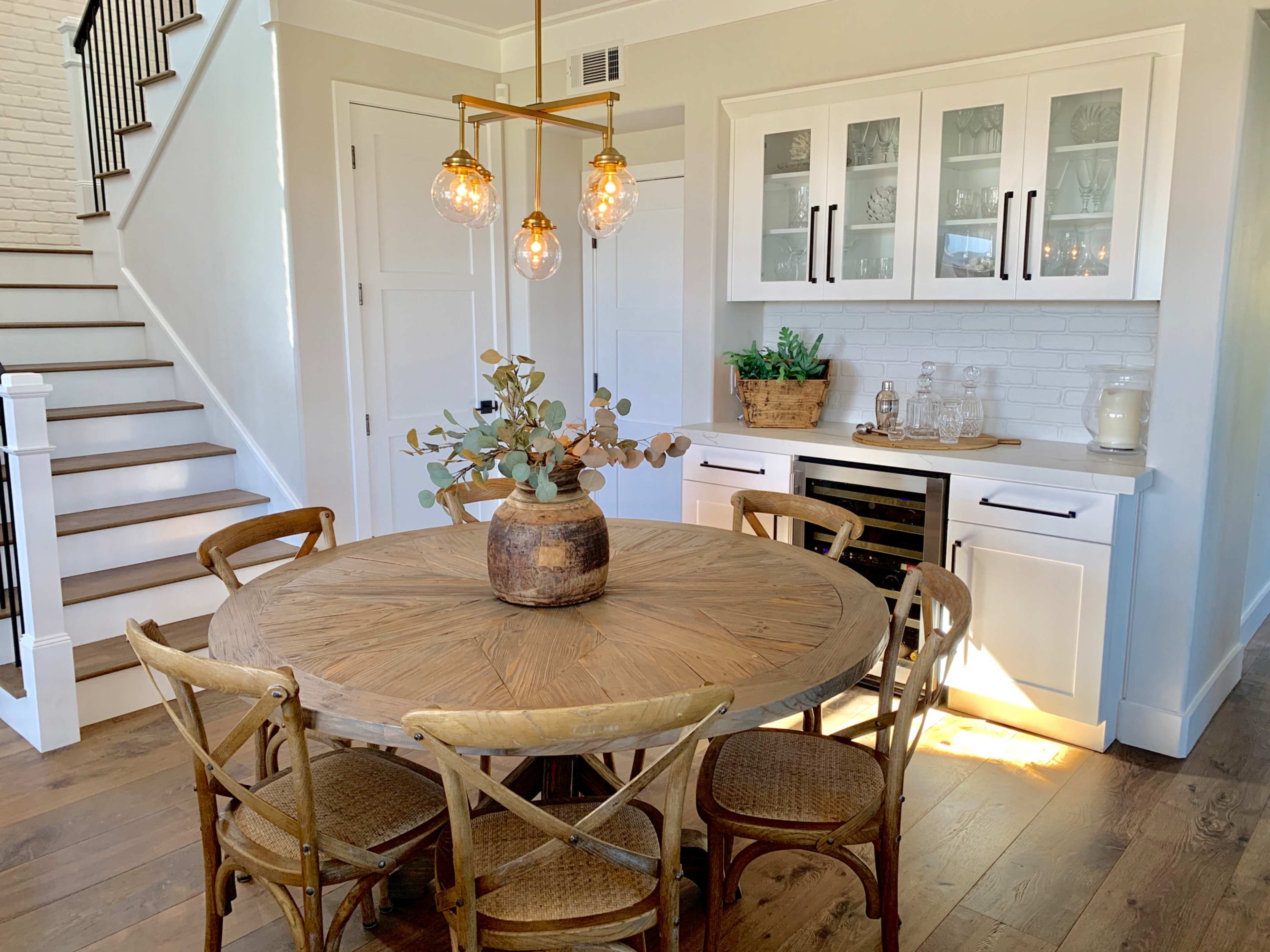 A round wooden dining table with six chairs is set in a well-lit kitchen area featuring modern cabinetry and a wine cooler.
