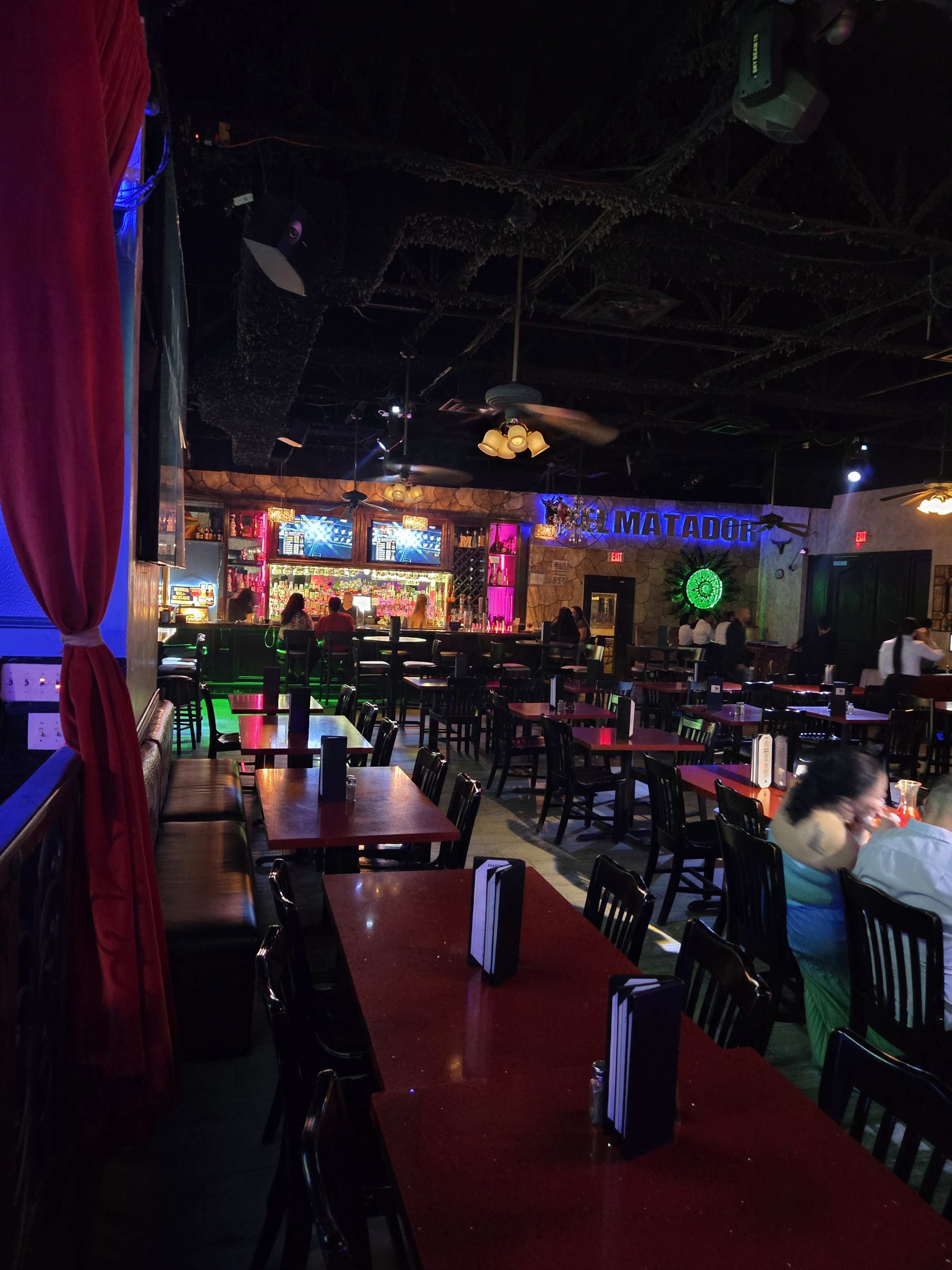 The image shows a dimly lit restaurant interior featuring rows of red-topped tables and black chairs, with a bar in the background illuminated by colorful lights.