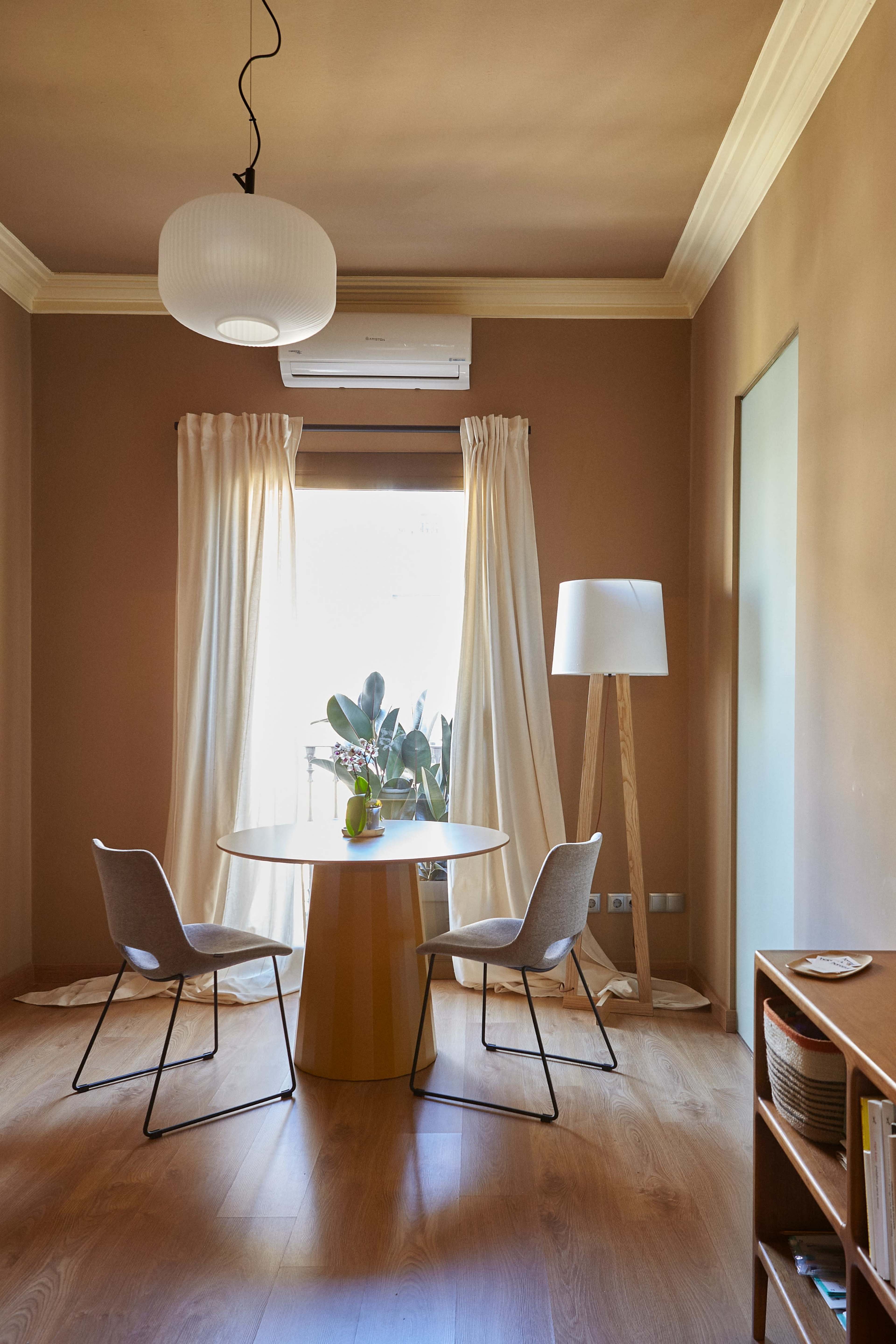 A round dining table with two chairs is positioned under a window, with a floor lamp nearby and curtains softly framing the view.