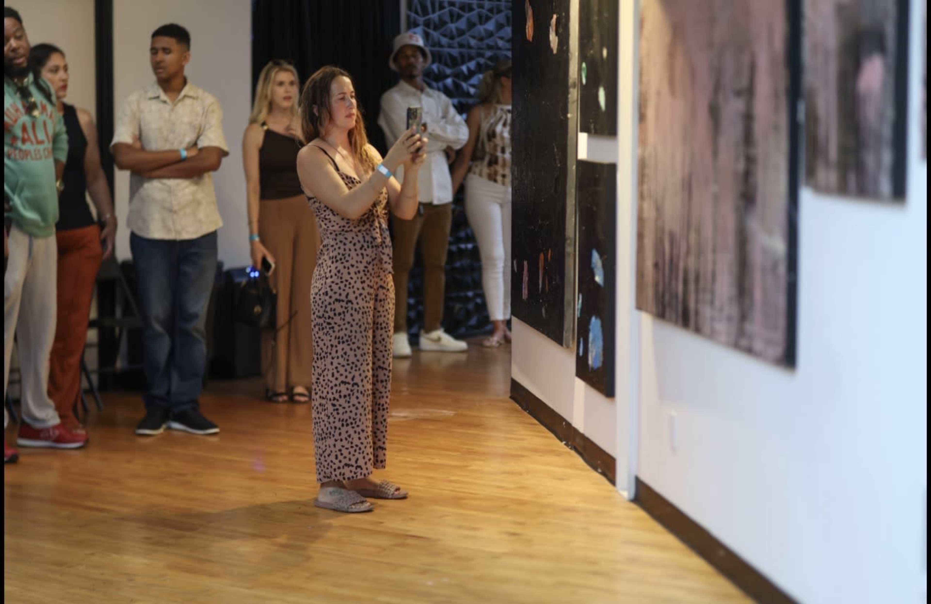 A woman in a brown patterned dress takes a photo of an art piece while a group of people watches in an art gallery.