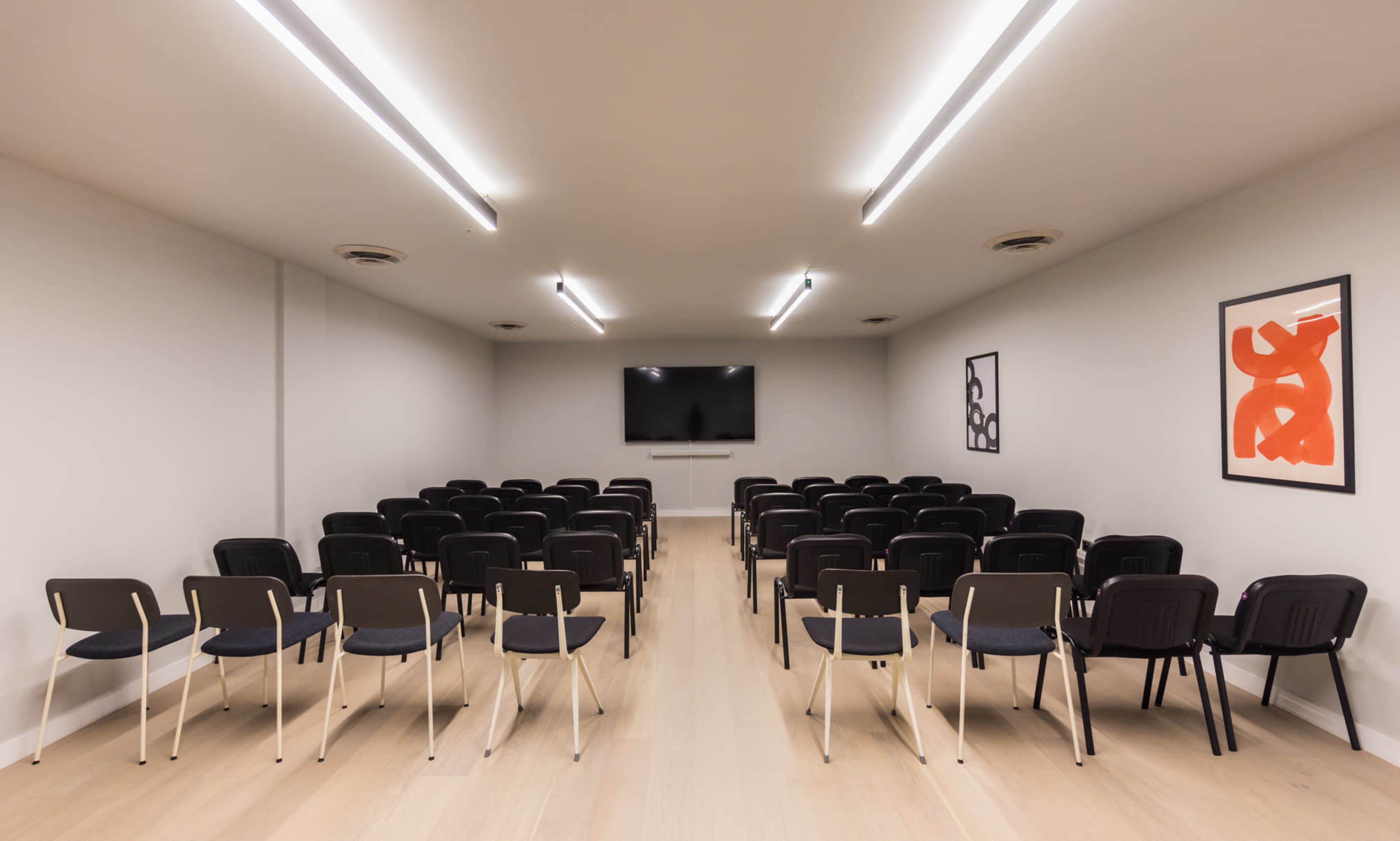The image shows a modern meeting room with rows of black chairs facing a wall-mounted television, illuminated by rectangular ceiling lights.
