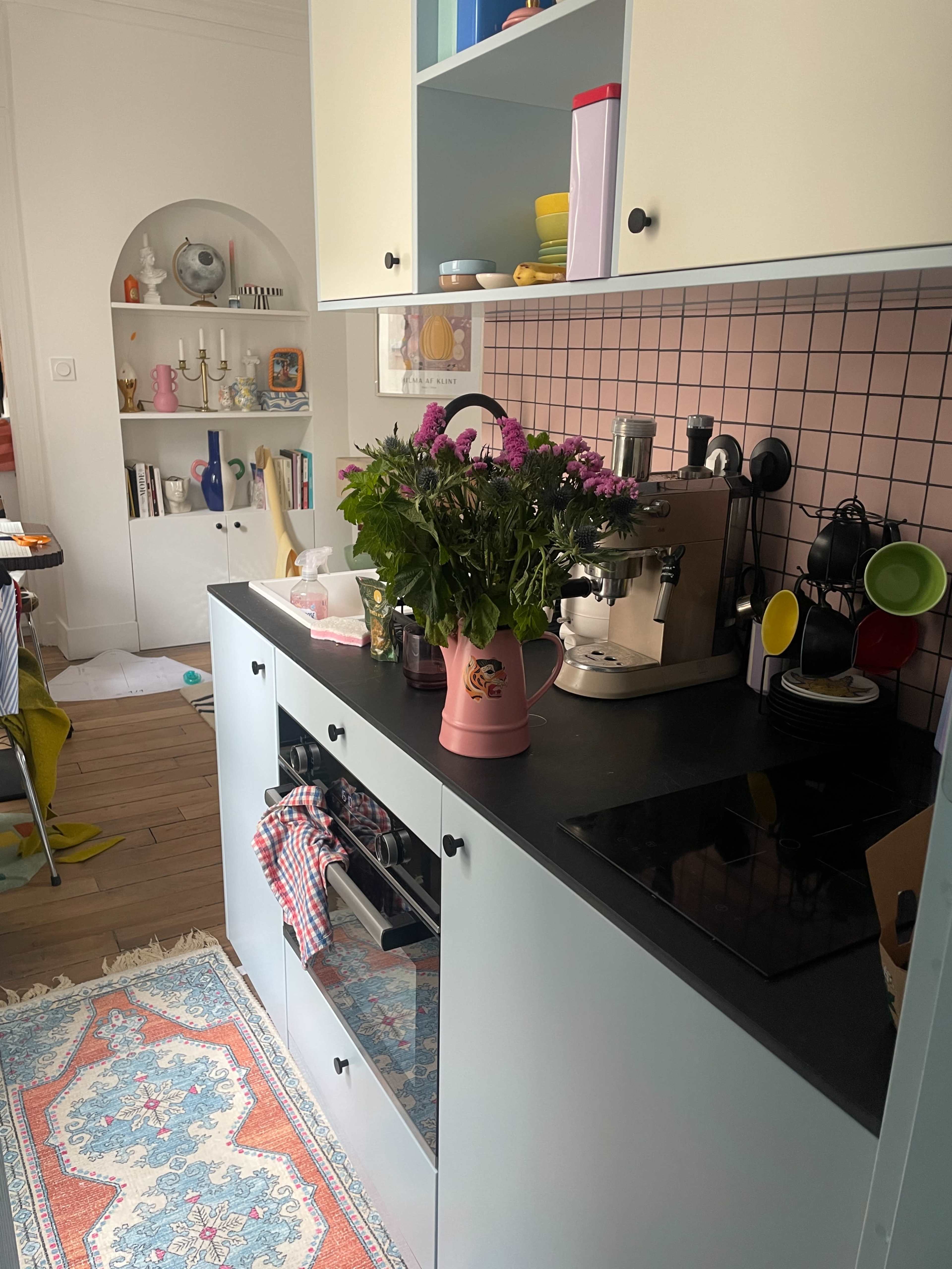 A compact kitchen features a colorful arrangement of dishes, a coffee machine, and a bouquet of flowers in a pitcher on the countertop.