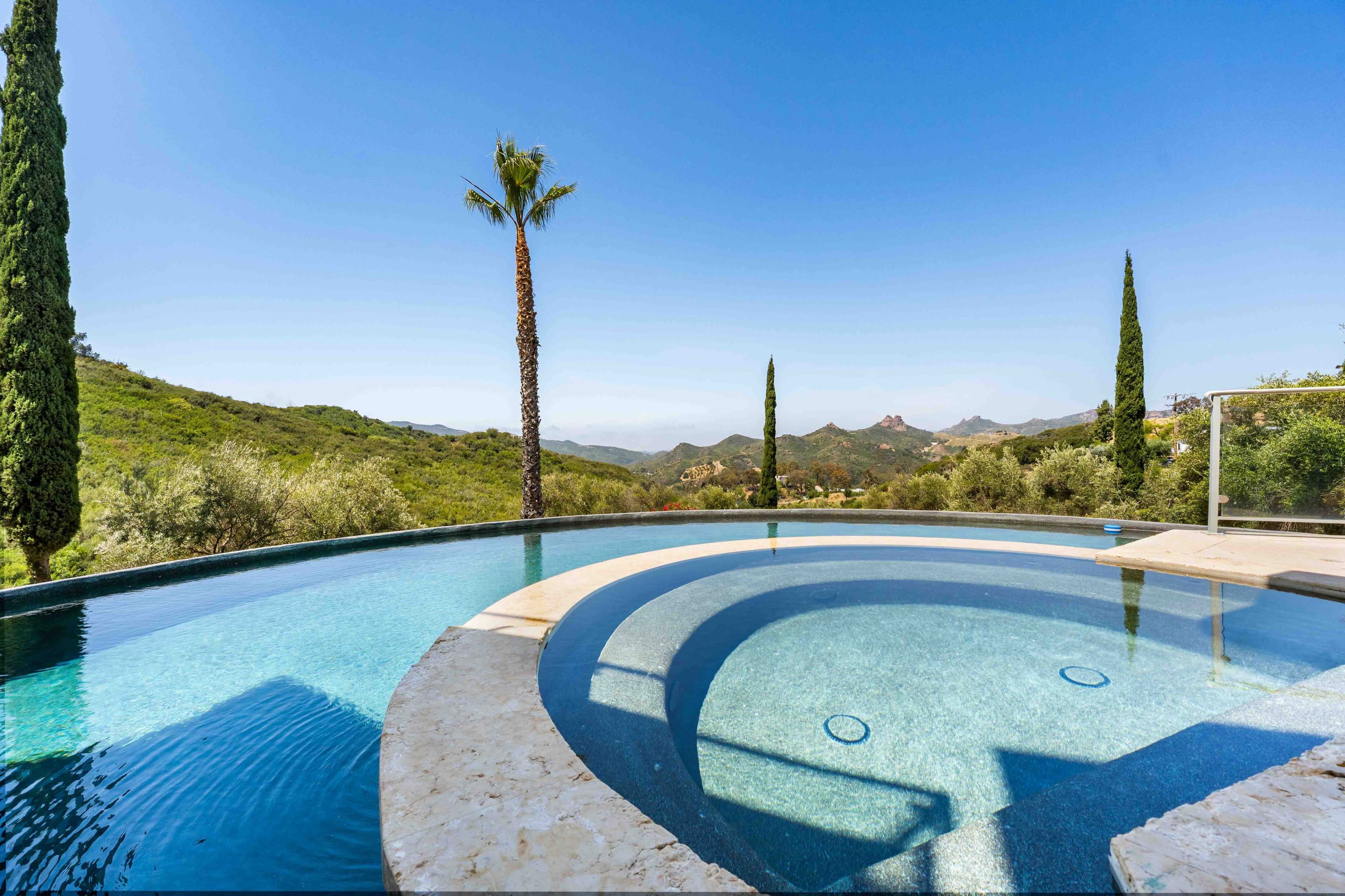 A swimming pool with a circular design is situated against a backdrop of rolling green hills and clear blue skies, with palm and cypress trees nearby.