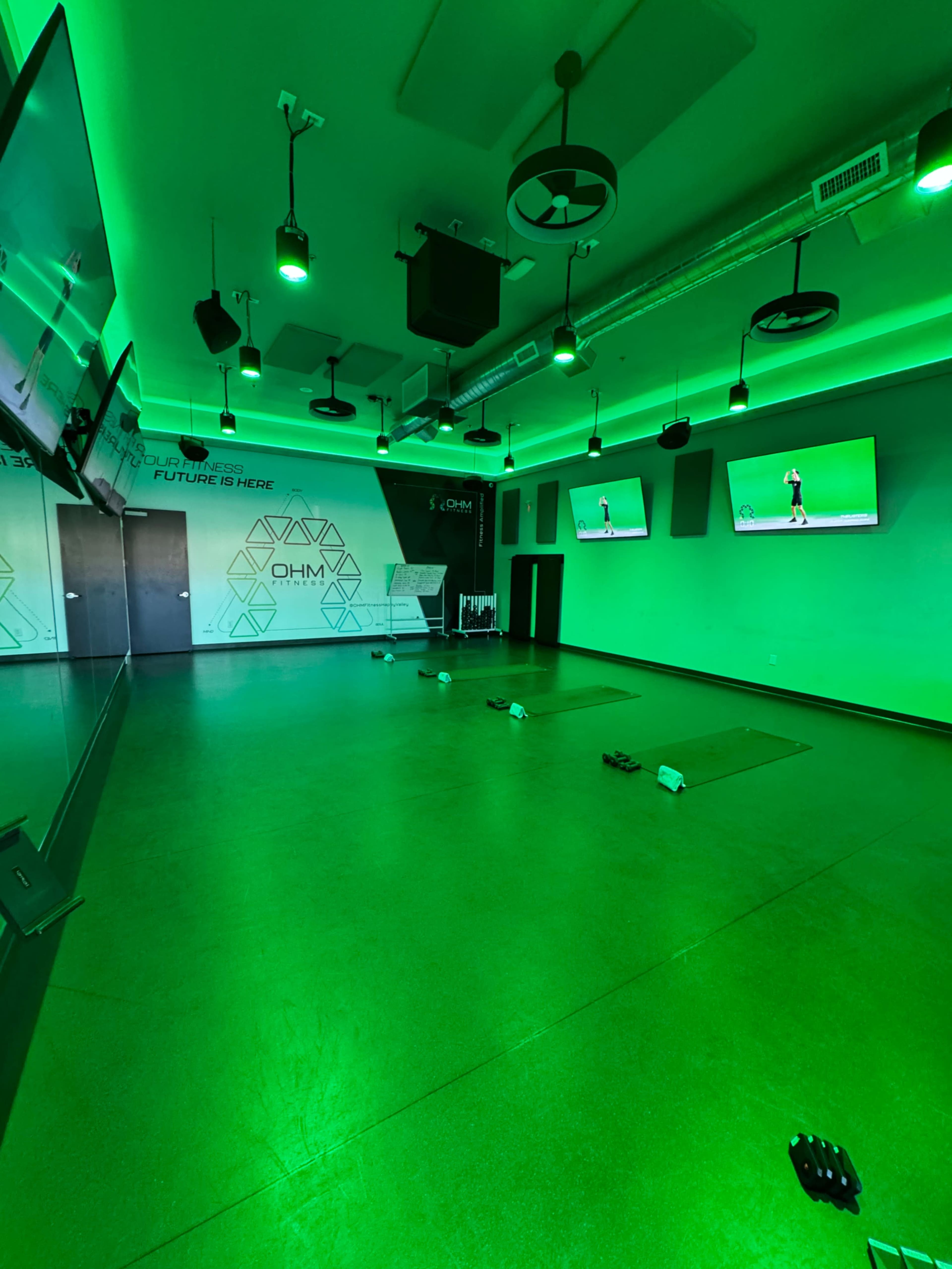 The image shows a modern fitness studio with green lighting, featuring large screens on the walls and exercise mats on the floor.