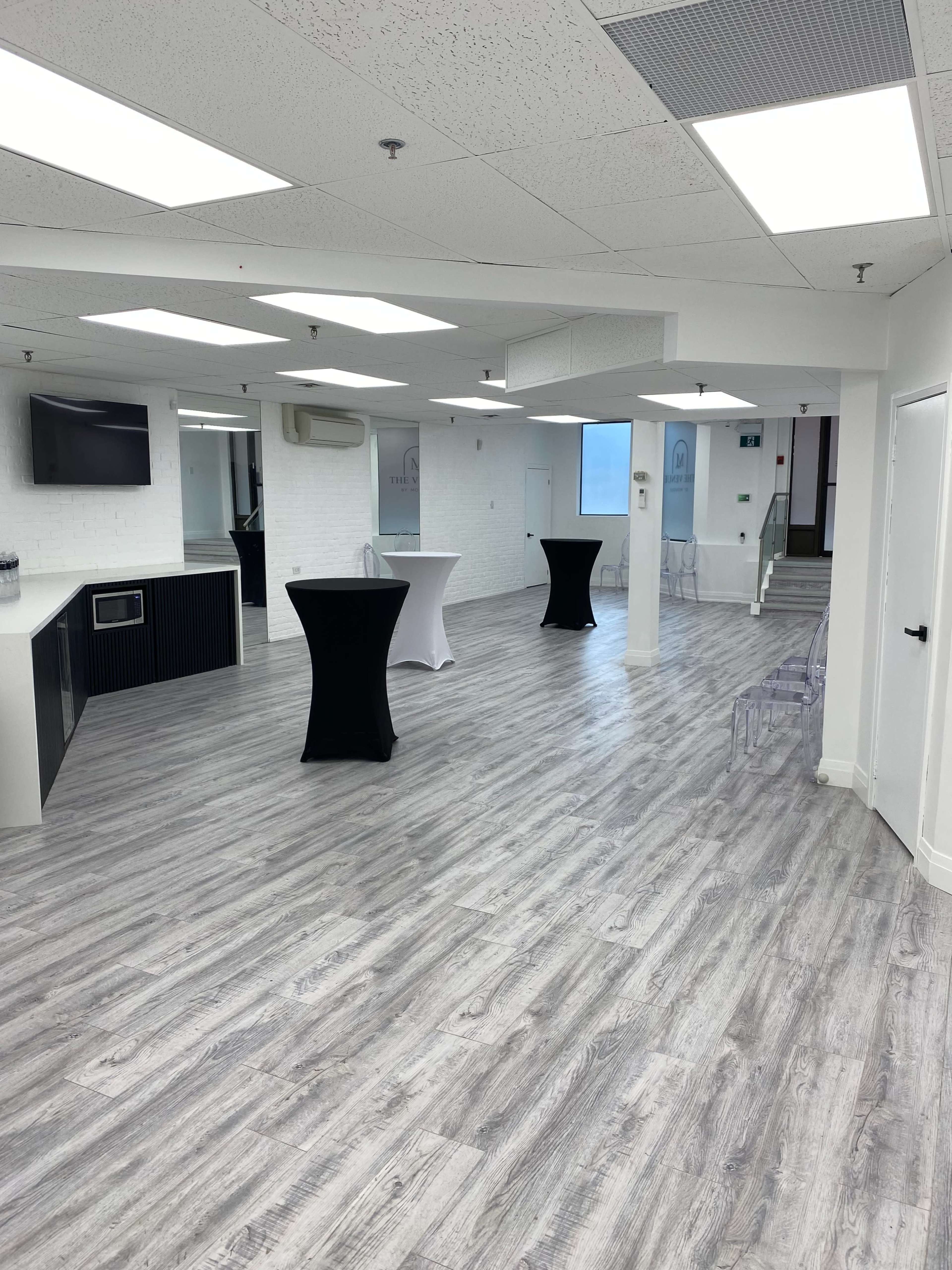 The image shows a spacious, empty room with wood-style flooring, featuring two black cocktail tables and a kitchenette area.
