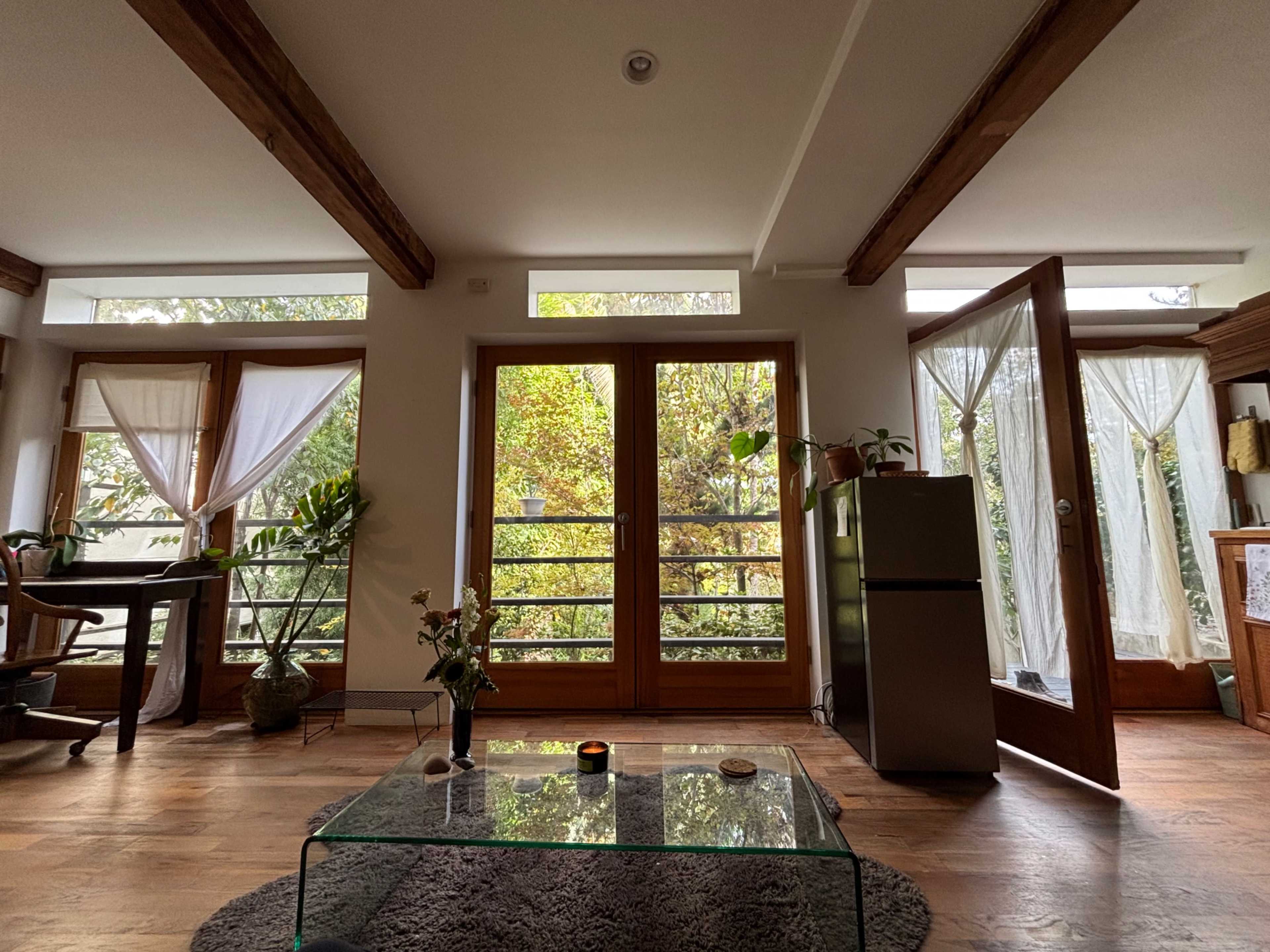 A bright living room features large glass doors opening to a lush garden, with wooden beams overhead and a glass coffee table in the center.