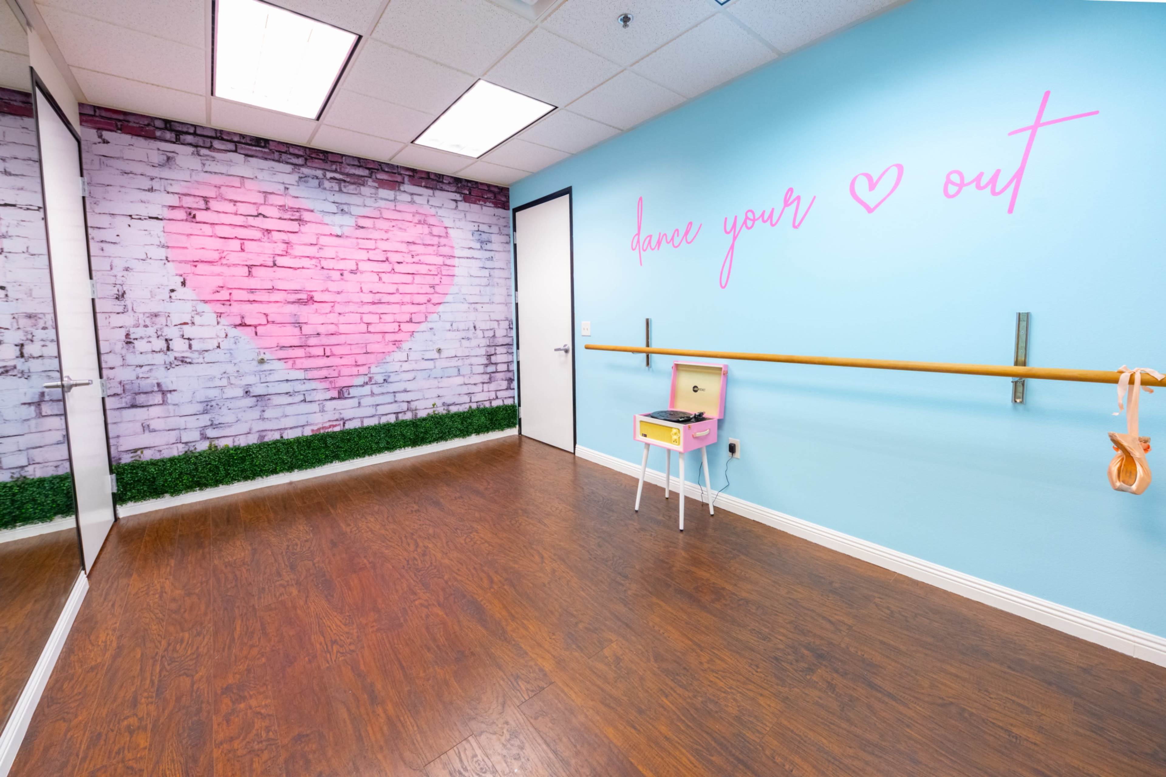 The image shows a dance studio with a pink heart mural on a brick wall, a ballet barre, and a small pink table with a decorative item.