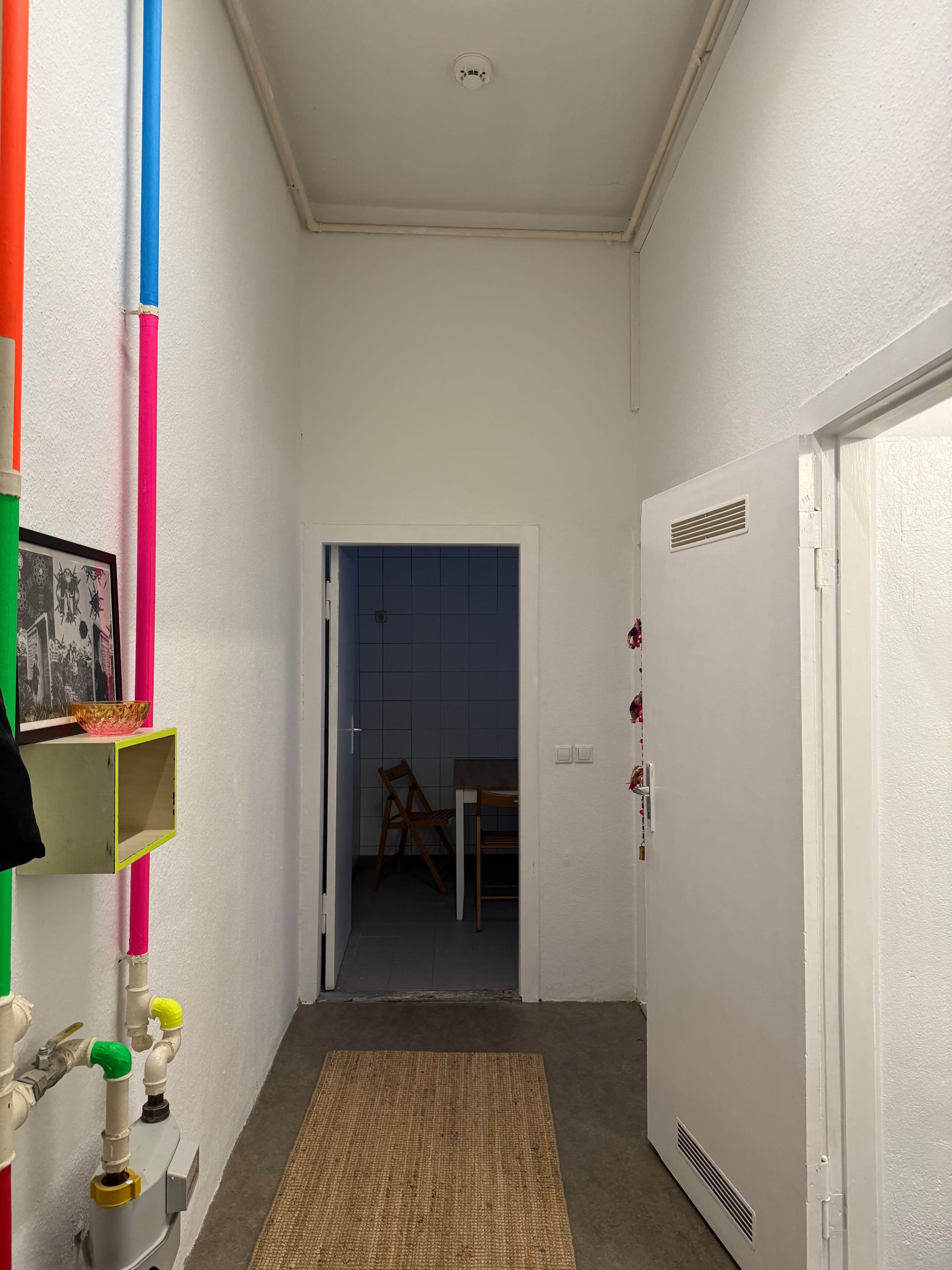 A narrow hallway features colorful pipes along the left wall and a door at the end leading to another room.