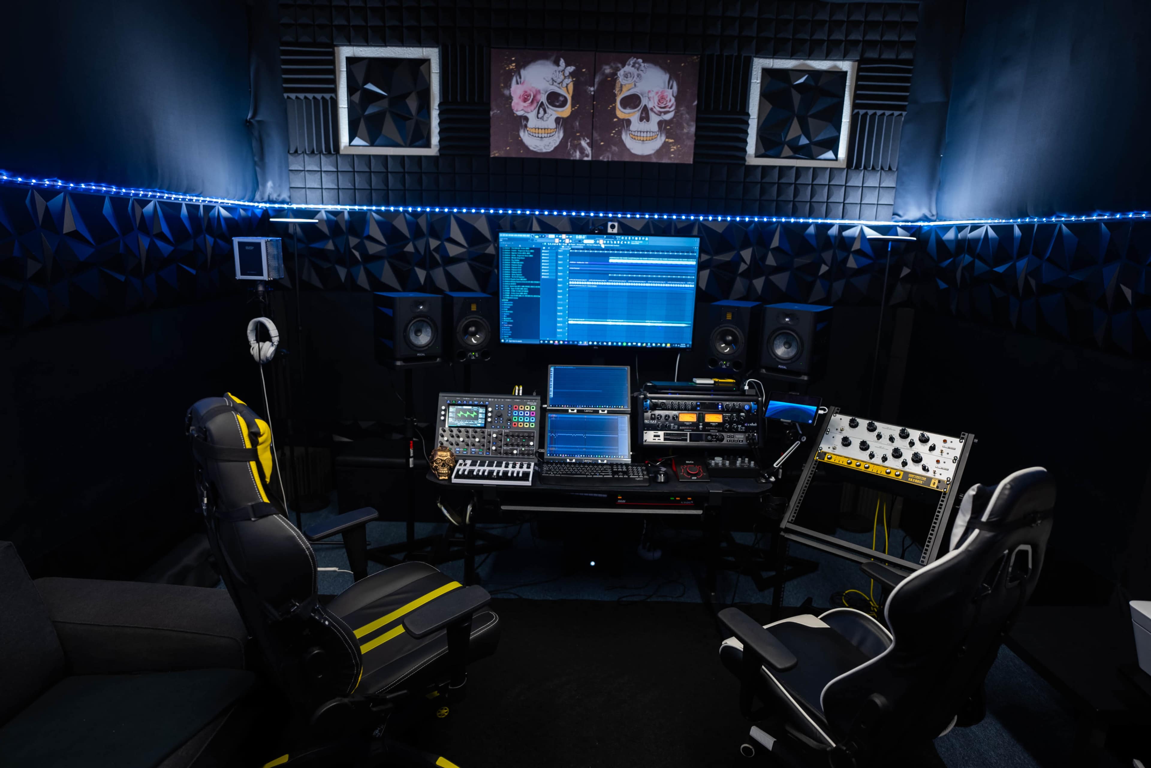 The image shows a music production studio with two chairs, multiple monitors, audio equipment, and soundproof wall panels.